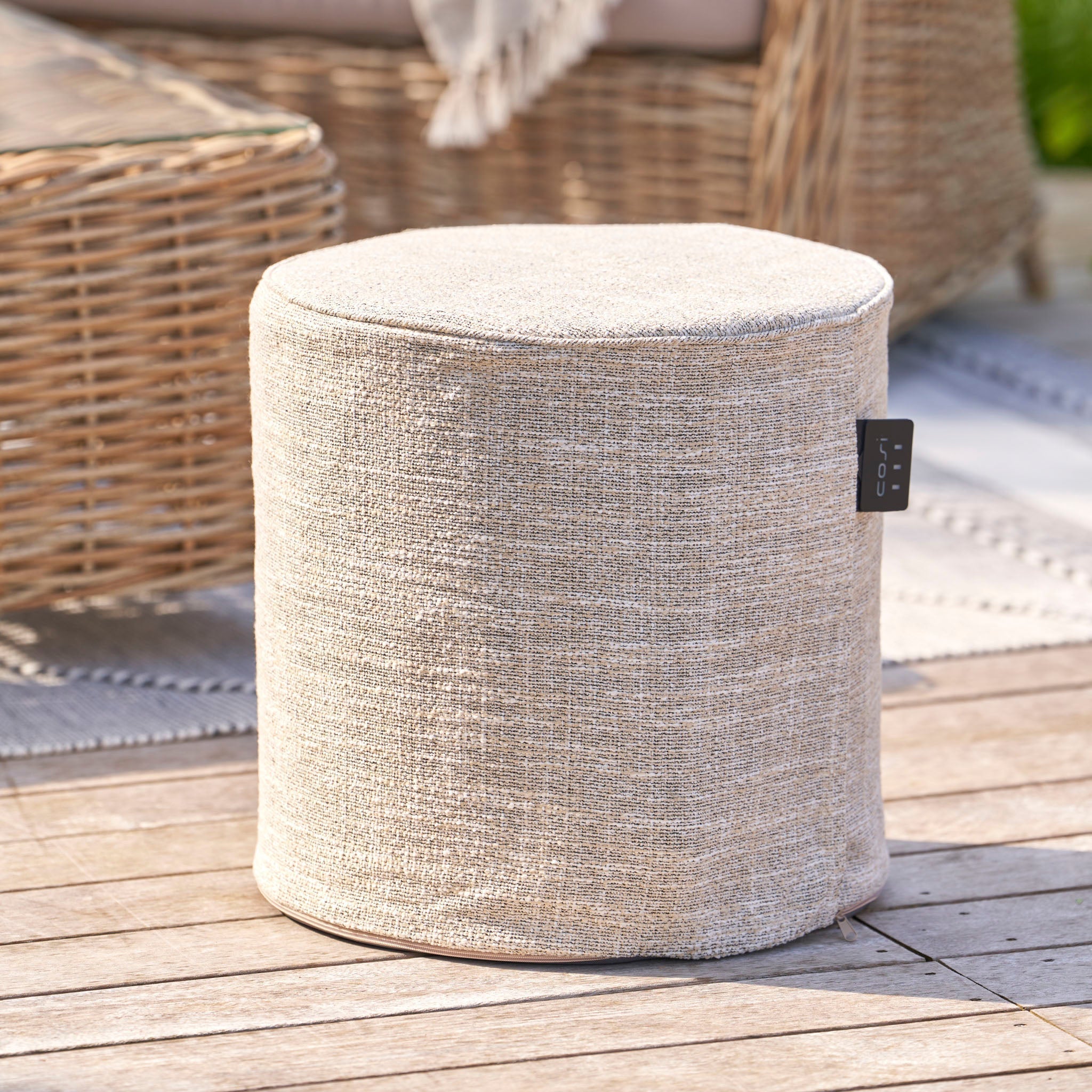 Beige woven ottoman on wooden deck with rattan furniture and rug in the background.