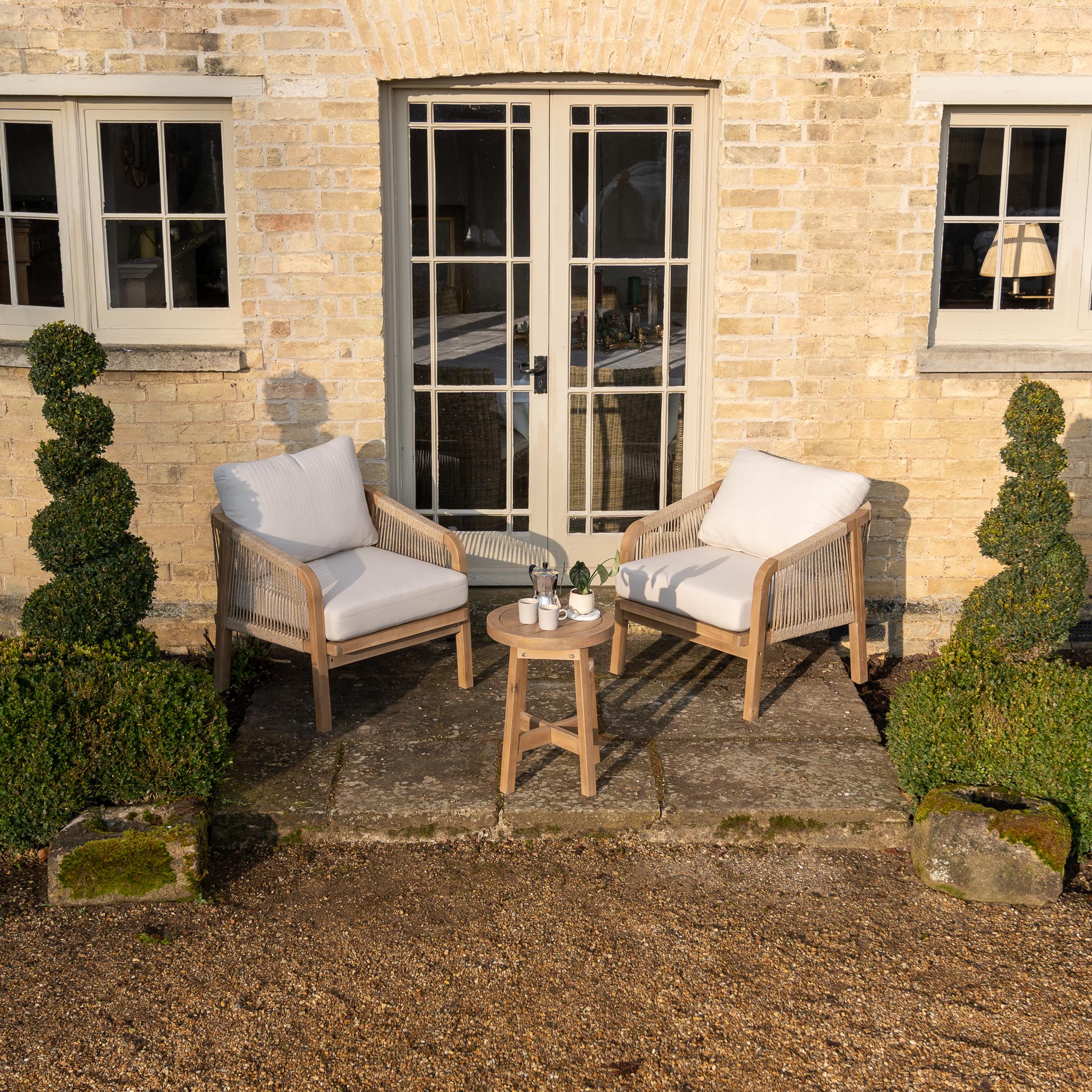 Two cushioned chairs and a small table with drinks, set outside a brick house with trimmed topiary shrubs.