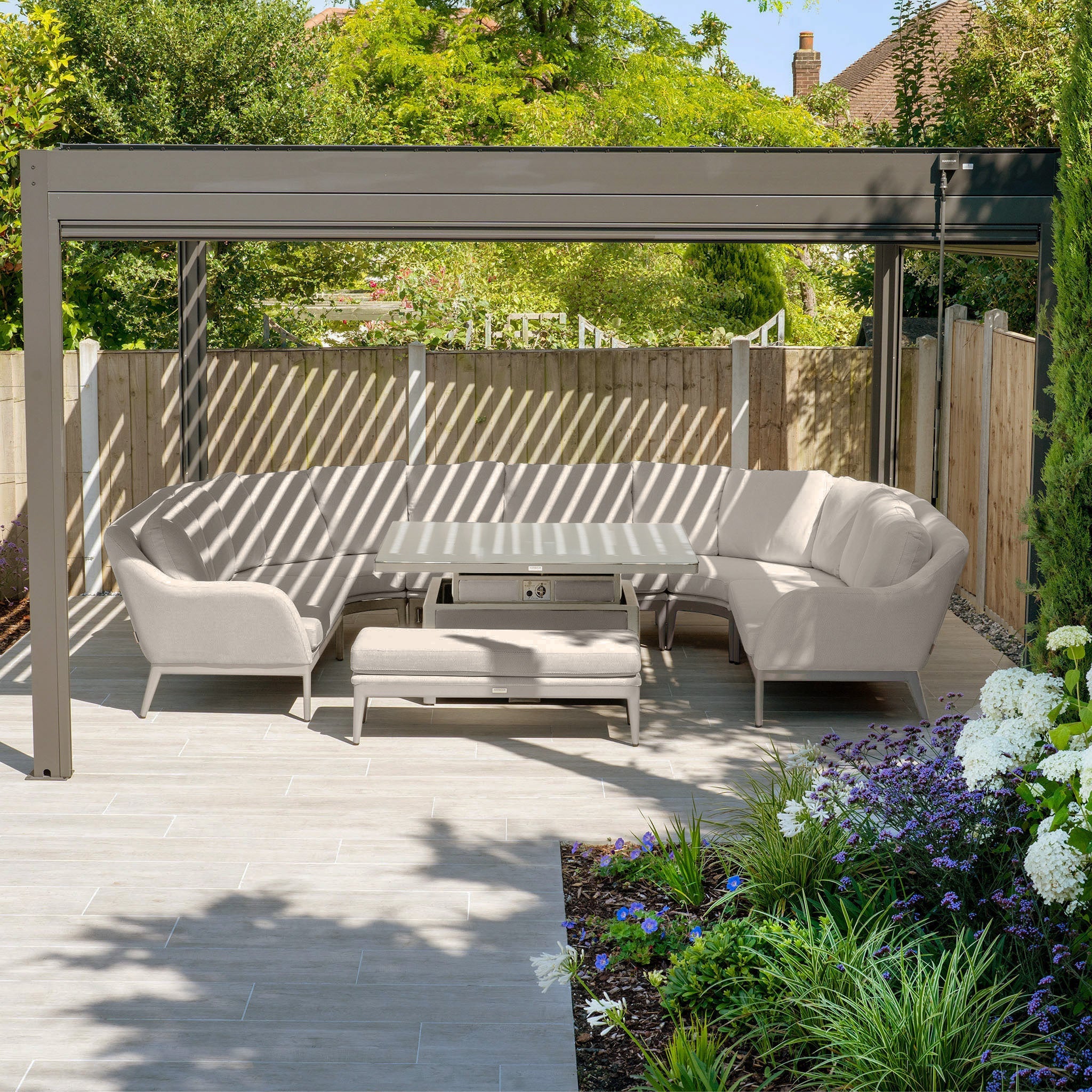 Outdoor seating area with a gray sectional sofa under a pergola, surrounded by a garden with purple and white flowers.