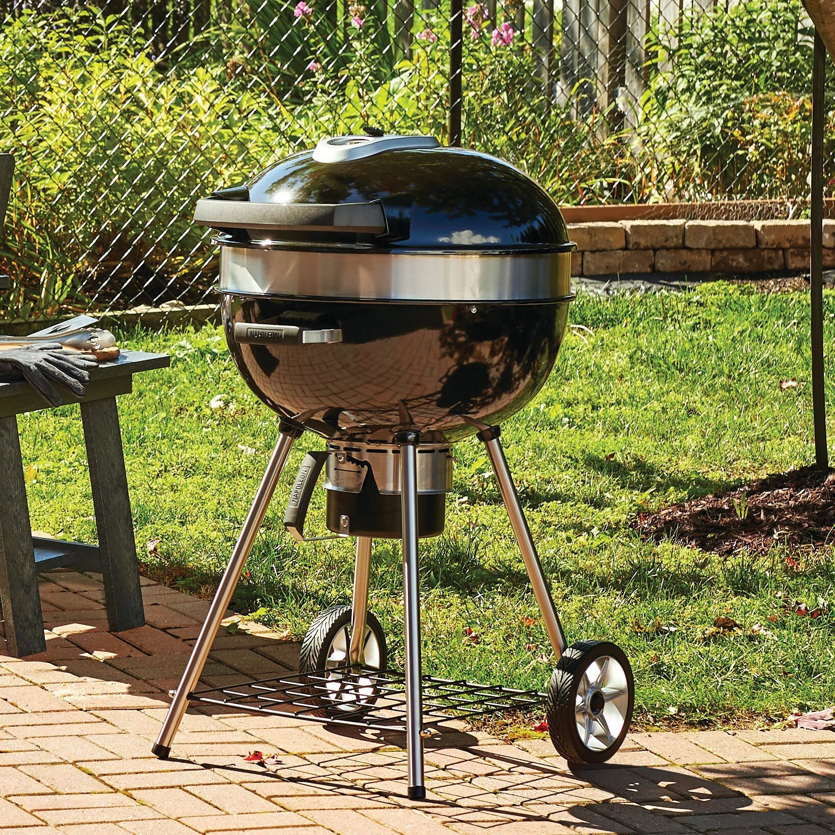 A black, round charcoal grill with a lid, standing on three metal legs, two of which have large wheels, and a lower shelf for storage.