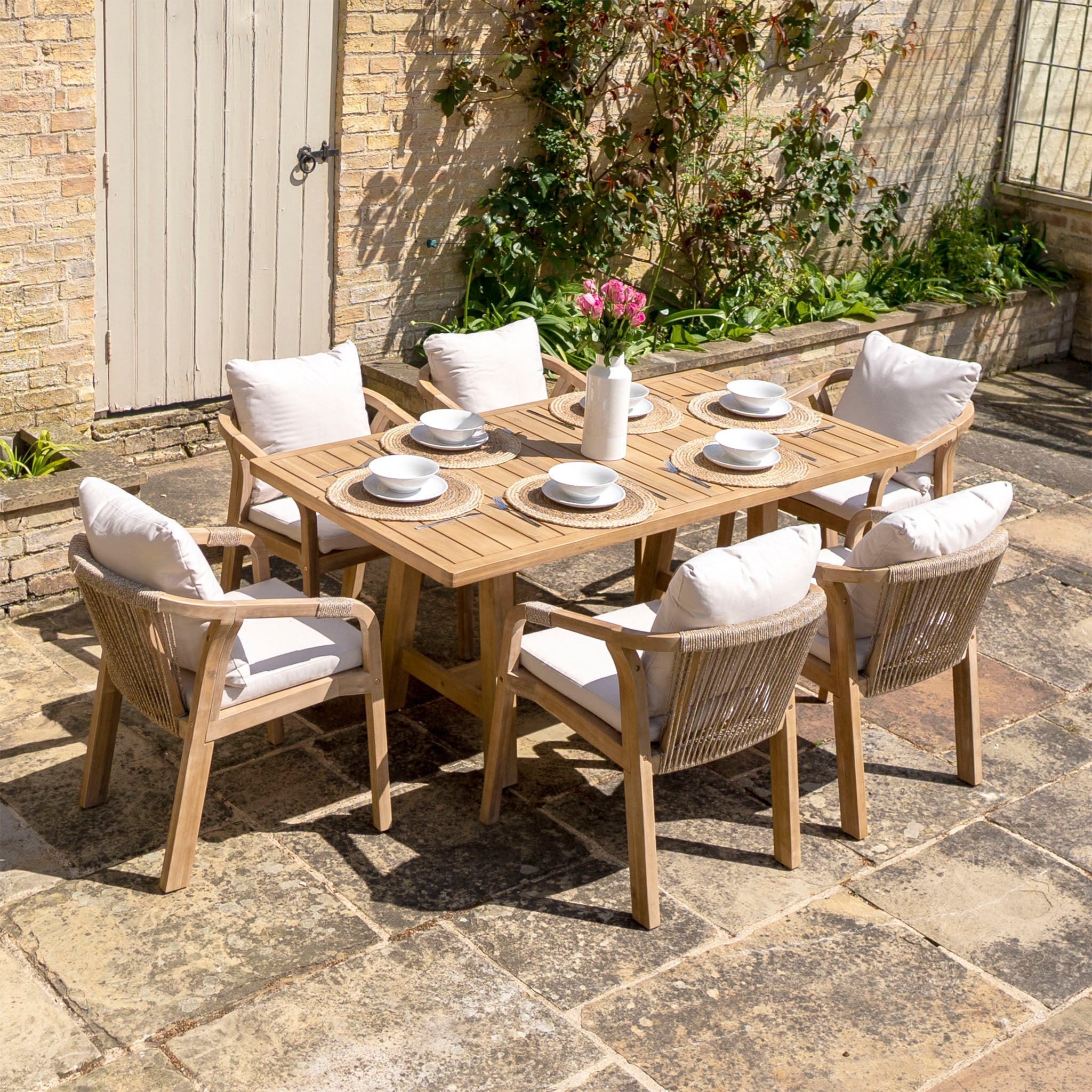 Outdoor wooden dining set with six cushioned chairs, set for a meal. Potted flowers on table; brick wall background.