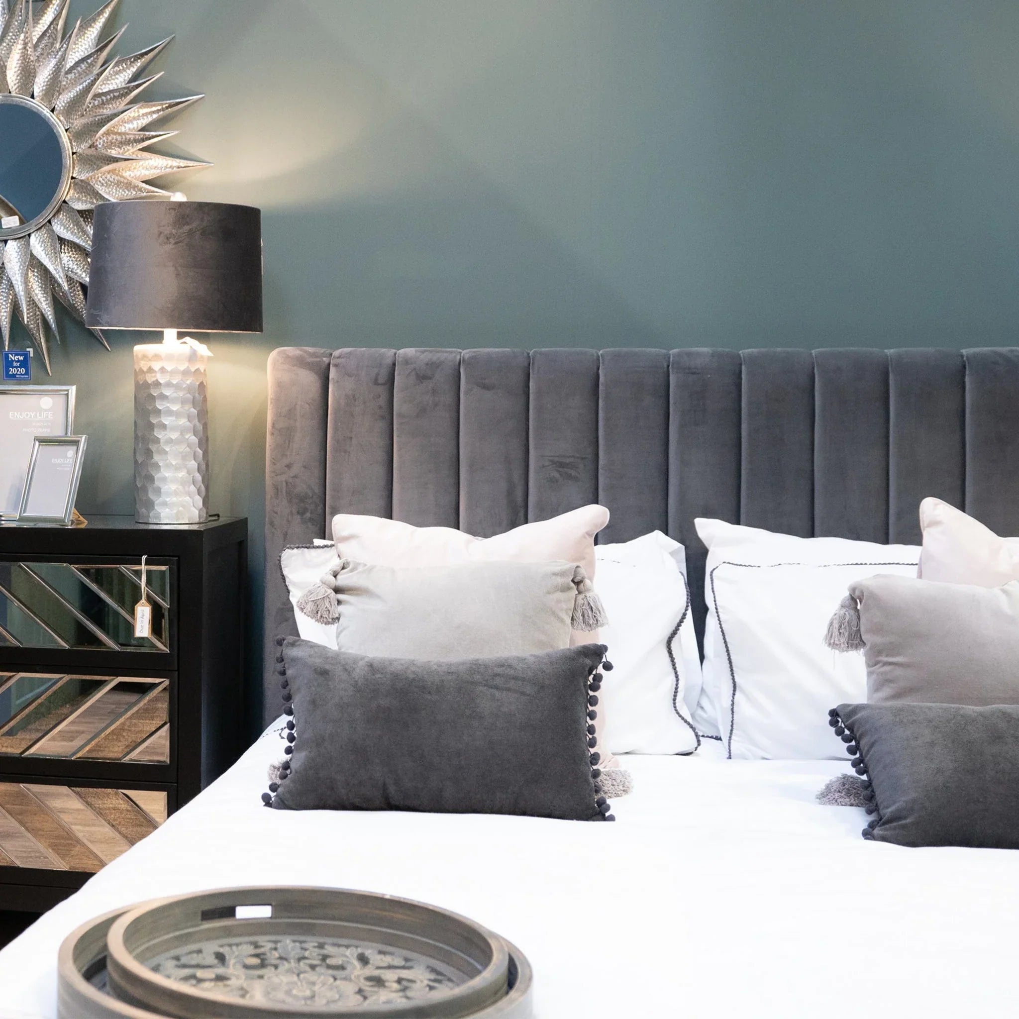 A modern bedroom features a grey upholstered headboard, decorative pillows, a bedside lamp, and a sunburst mirror.