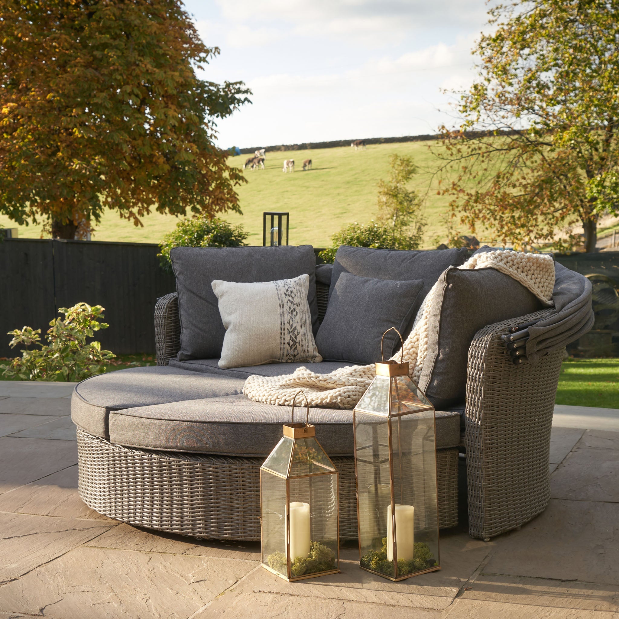 Cozy outdoor seating with gray cushions, throw blanket, and lanterns on a patio, overlooking a pastoral landscape.