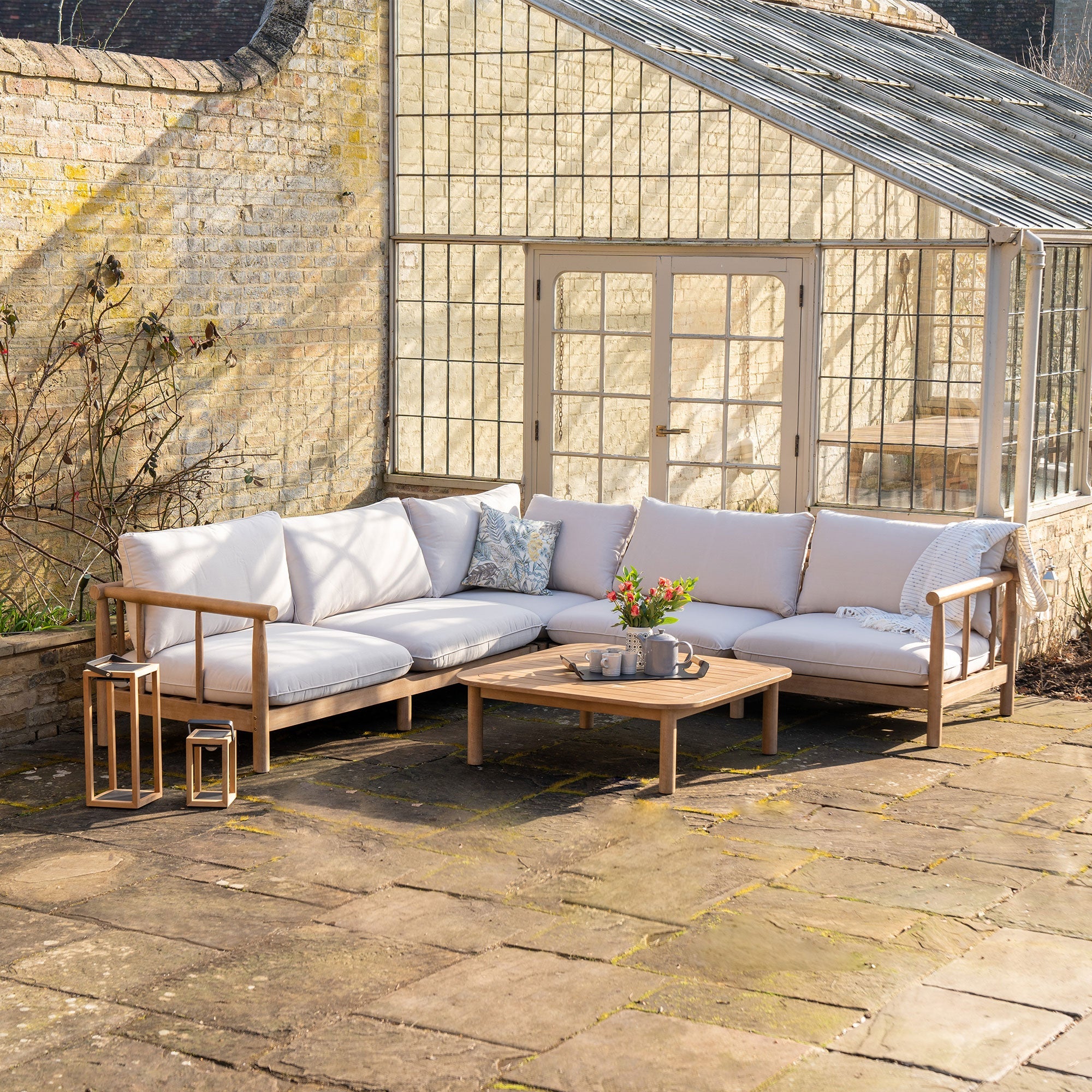 Outdoor patio with a wooden corner sofa, cushions, a coffee table, and lanterns. Glasshouse in the background.