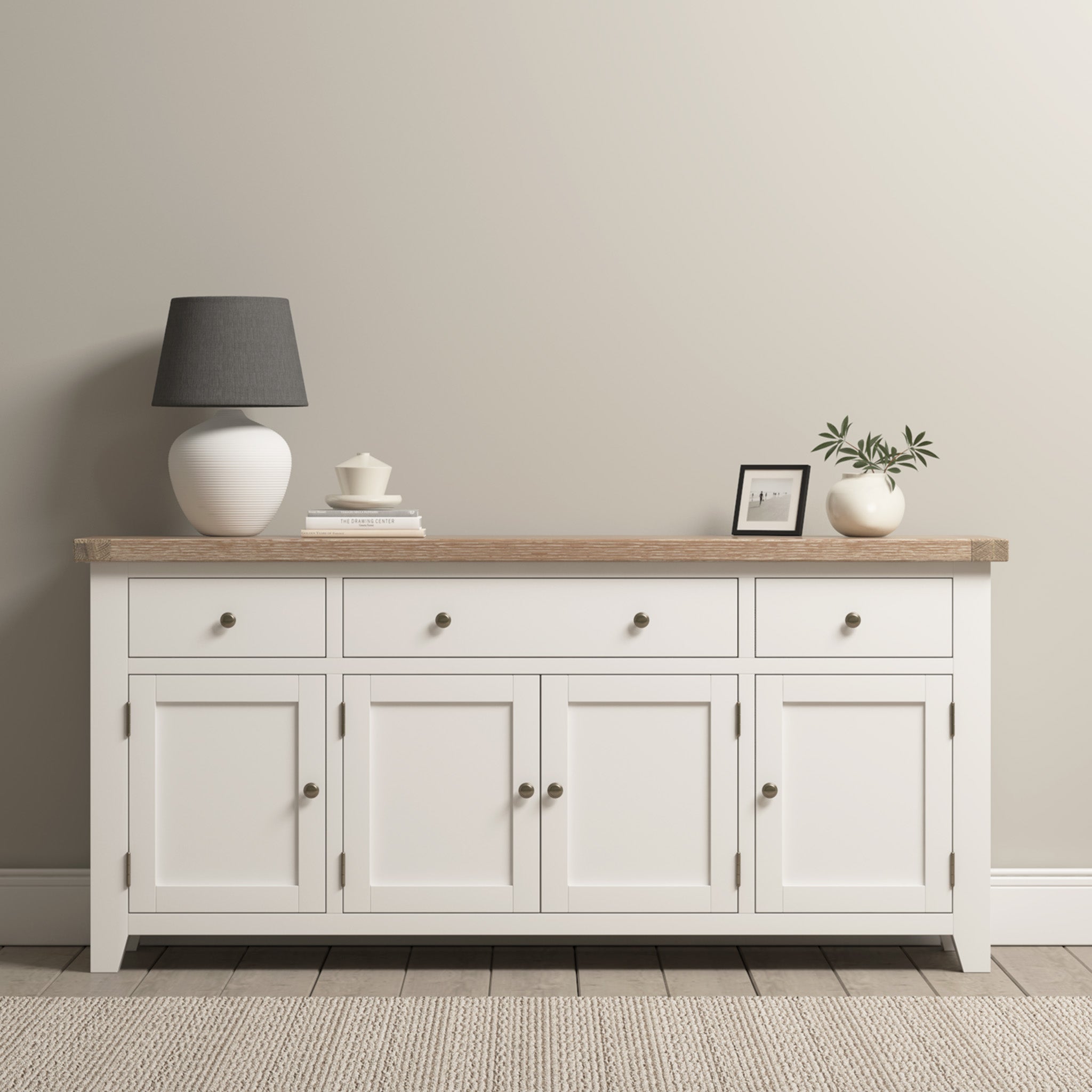 White sideboard with a wooden top, lamp, books, small framed photo, and potted plant on a neutral wall background.