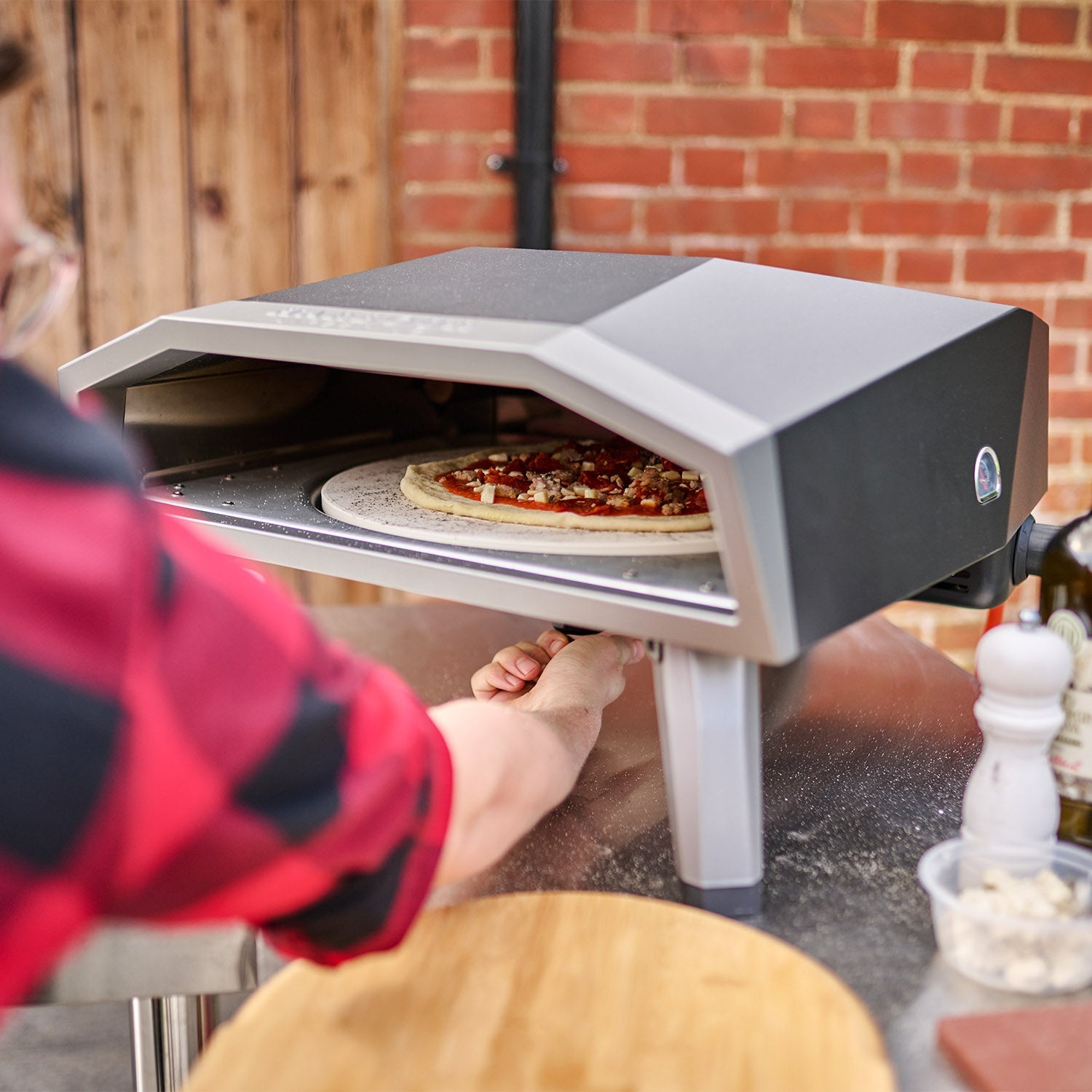 Person placing a pizza in an outdoor pizza oven on a table, next to olive oil and a pepper mill.