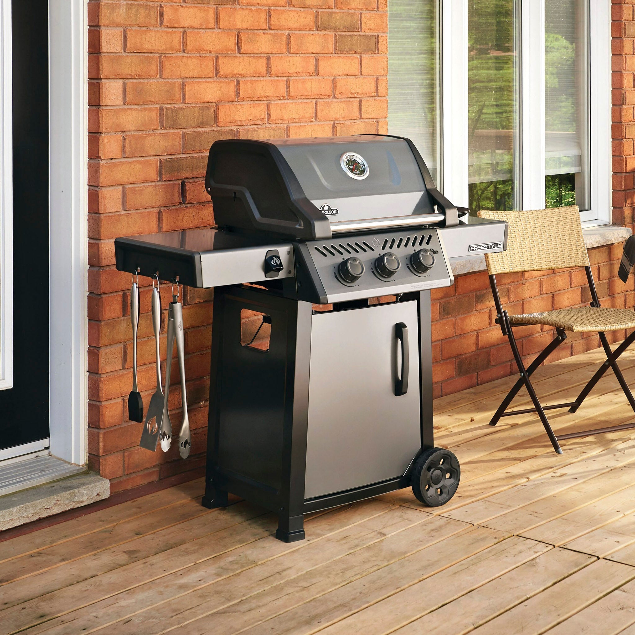 A stainless steel barbecue grill with utensils hangs on its side, placed on a wooden deck near a brick wall and chair.