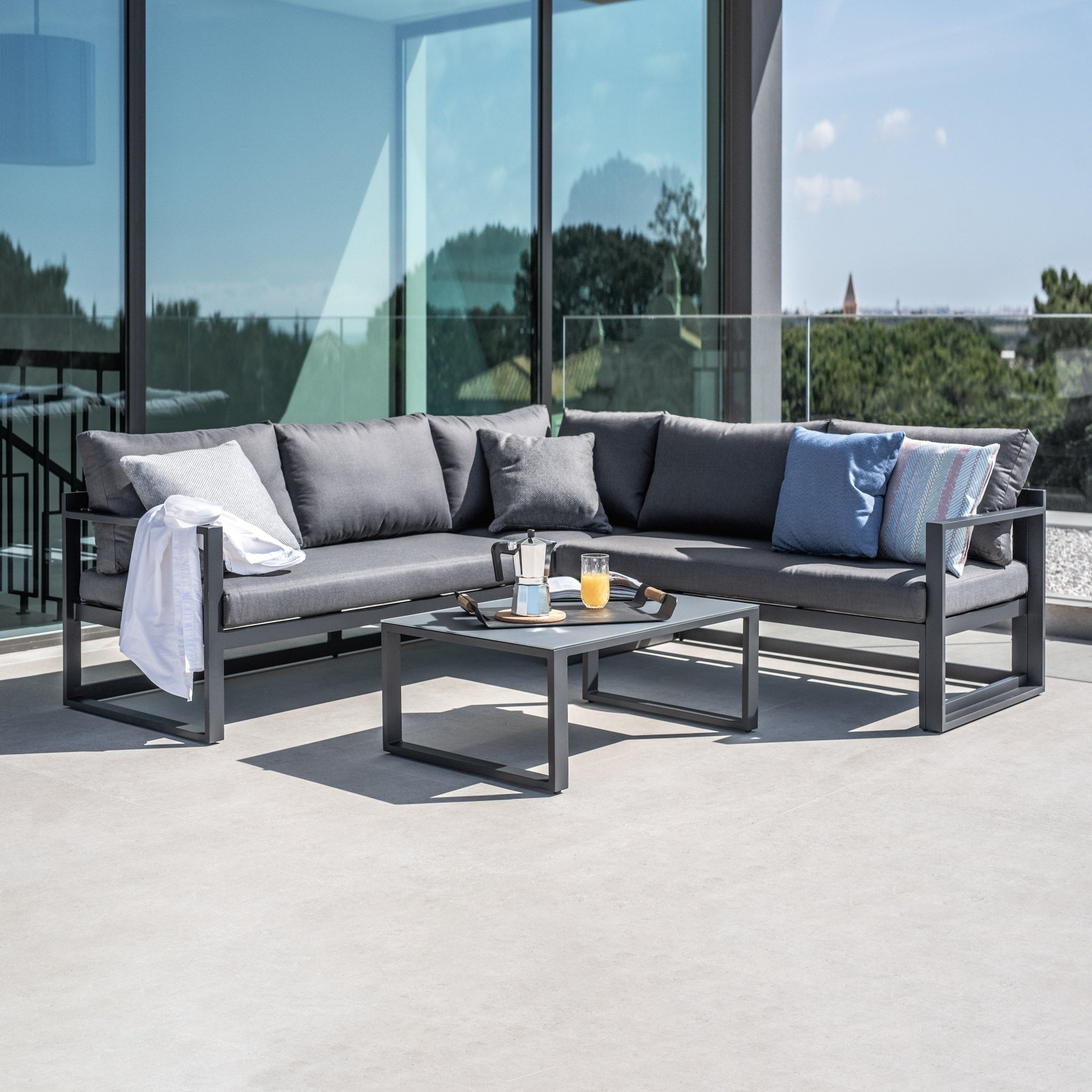 Modern patio with gray sectional sofa, coffee table, pillows, and drinks. Glass railing with a scenic view in the background.