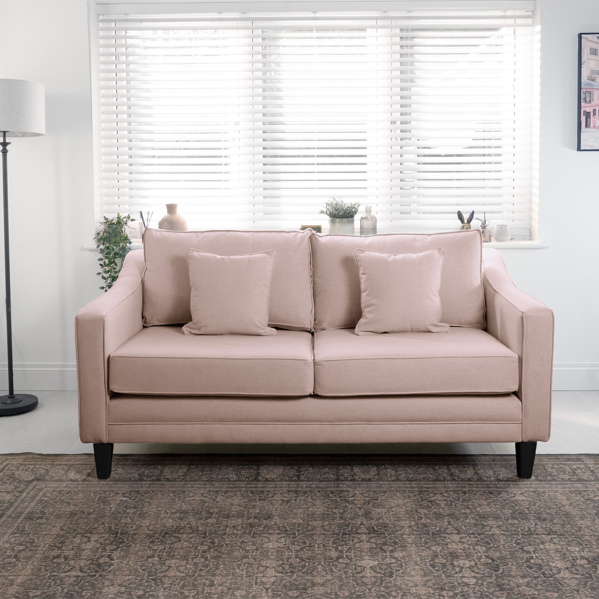 A light pink sofa with pillows in front of a window with white blinds, next to a floor lamp and indoor plants.