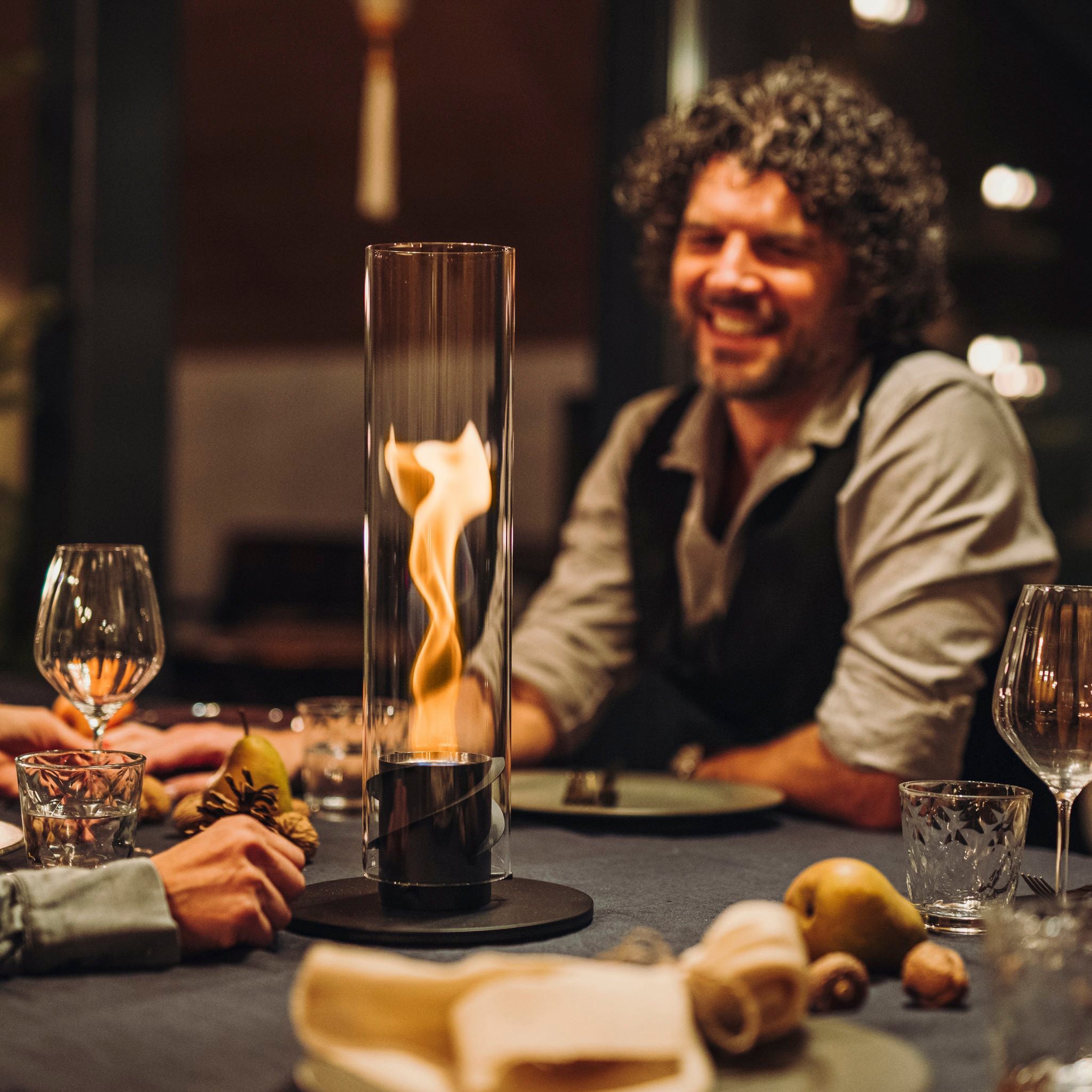 A person with curly hair smiles at a dining table adorned with the stylish Höfats SPIN 90 Table Top Fire in Black and an elegant table setting.