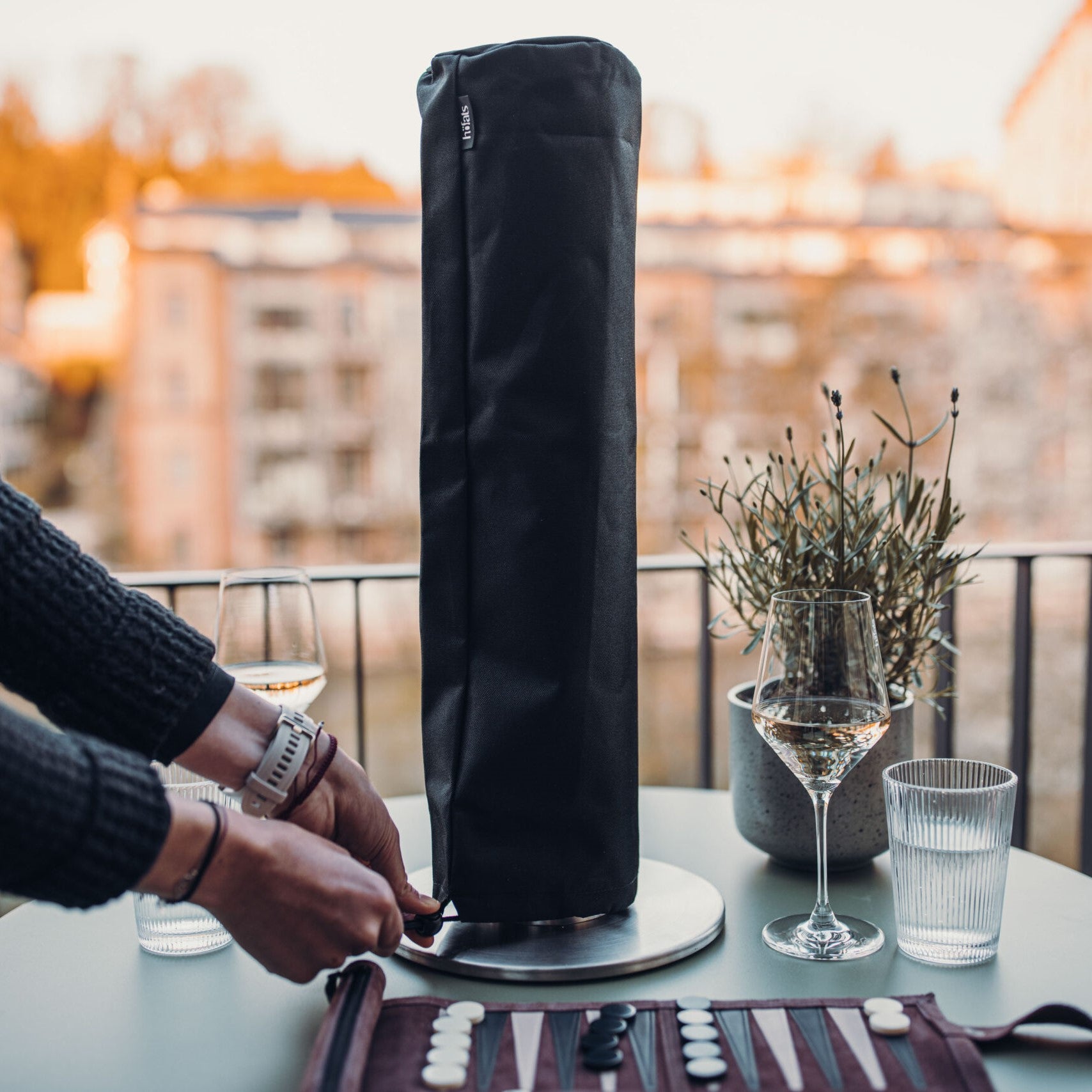 A person adjusts the Höfats SPIN 120 & 1200 Cover on a tabletop appliance on a balcony, ensuring its secure fastening while a wine glass, glass of water, and game board sit nearby.