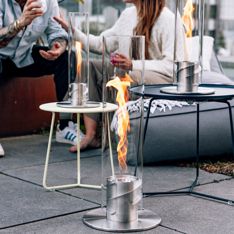 Two people sit around the Höfats SPIN 1500 Tabletop Fireplace Silver on a patio, enjoying drinks and conversation as the bioethanol fuels a mesmerizing flame whirl.