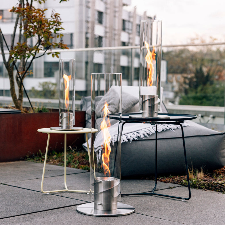 Three Höfats SPIN 1500 Tabletop Fireplaces in silver stand elegantly on the patio, their decorative glass cylinders enhancing the scene. Buildings and trees provide a picturesque backdrop on this cloudy day, while a mesmerizing flame whirl dances inside each cylinder.