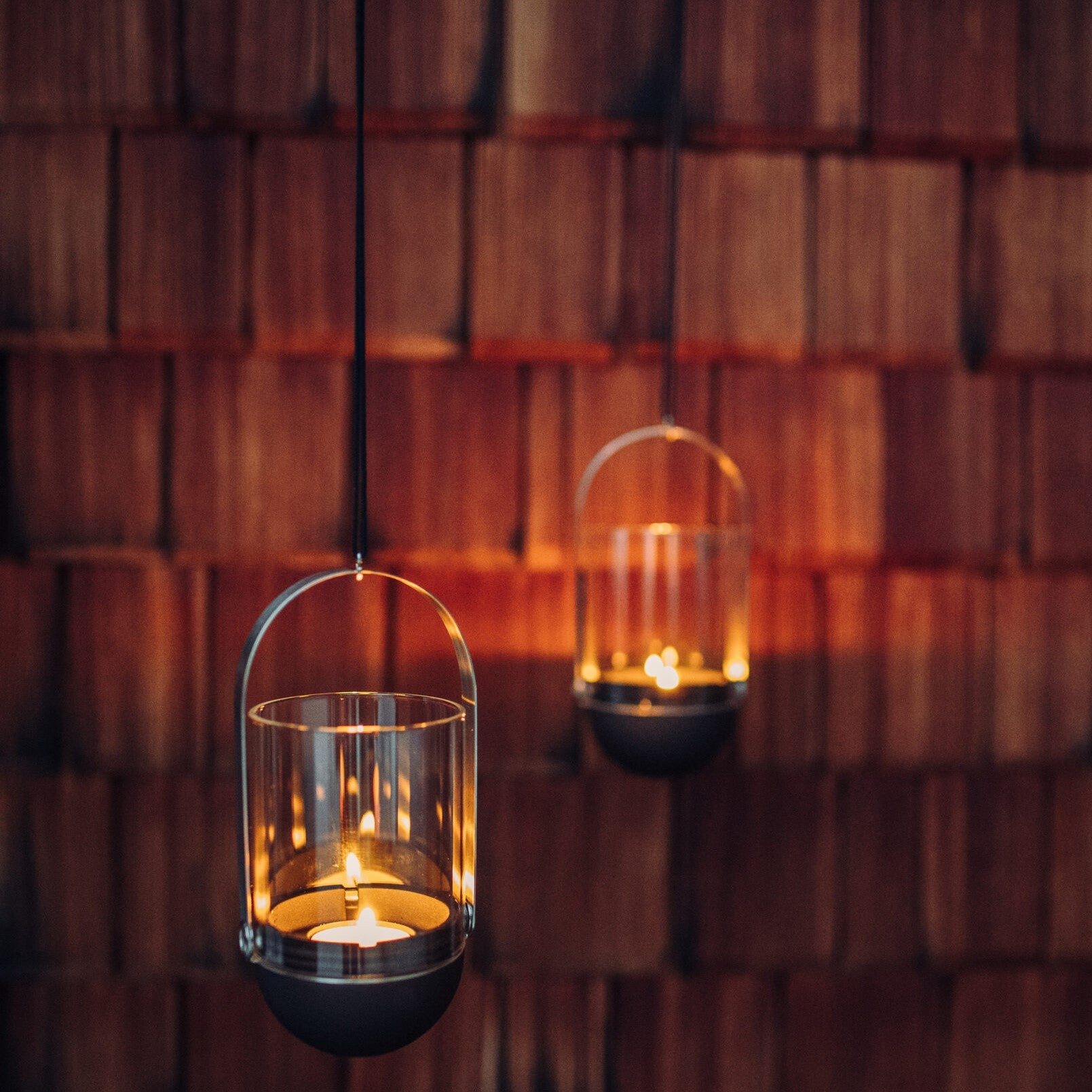 Two Höfats GRAVITY CANDLE Hangers, with their brushed solid stainless steel finish gleaming subtly in the light, cast a warm glow against a wooden, shingled wall background.