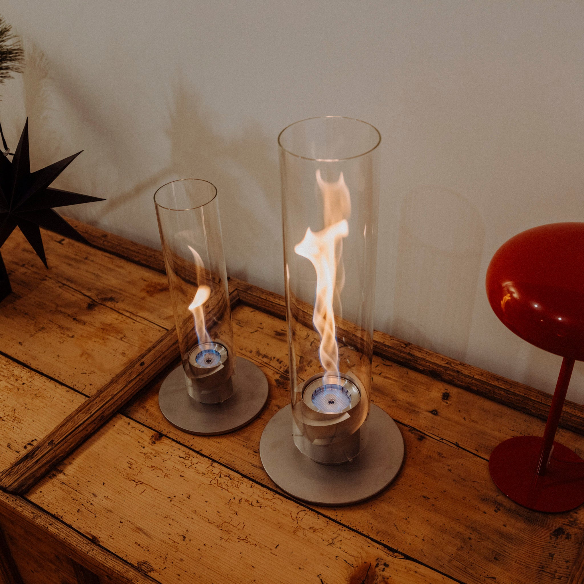 Two Höfats SPIN 900 Tabletop Fireplaces in grey, featuring burning flames encapsulated in glass cylinders and fueled by bioethanol, sit on a wooden surface next to a red lamp.