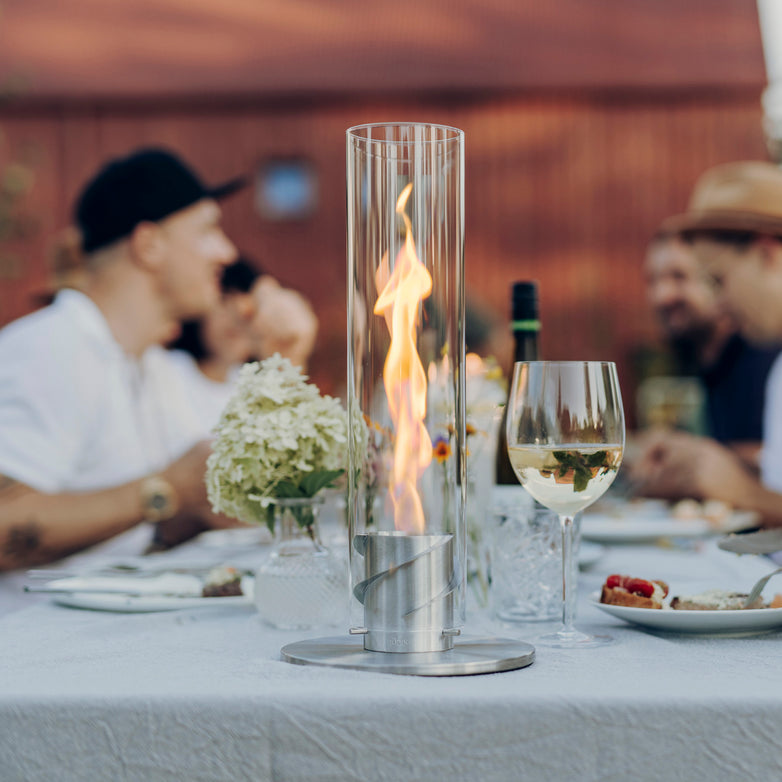 A table set with food and wine features a Höfats SPIN 90 Table Top Fire in Silver, showcasing a decorative glass cylinder fireplace. In the background, people socialize, adding warmth to the scene.