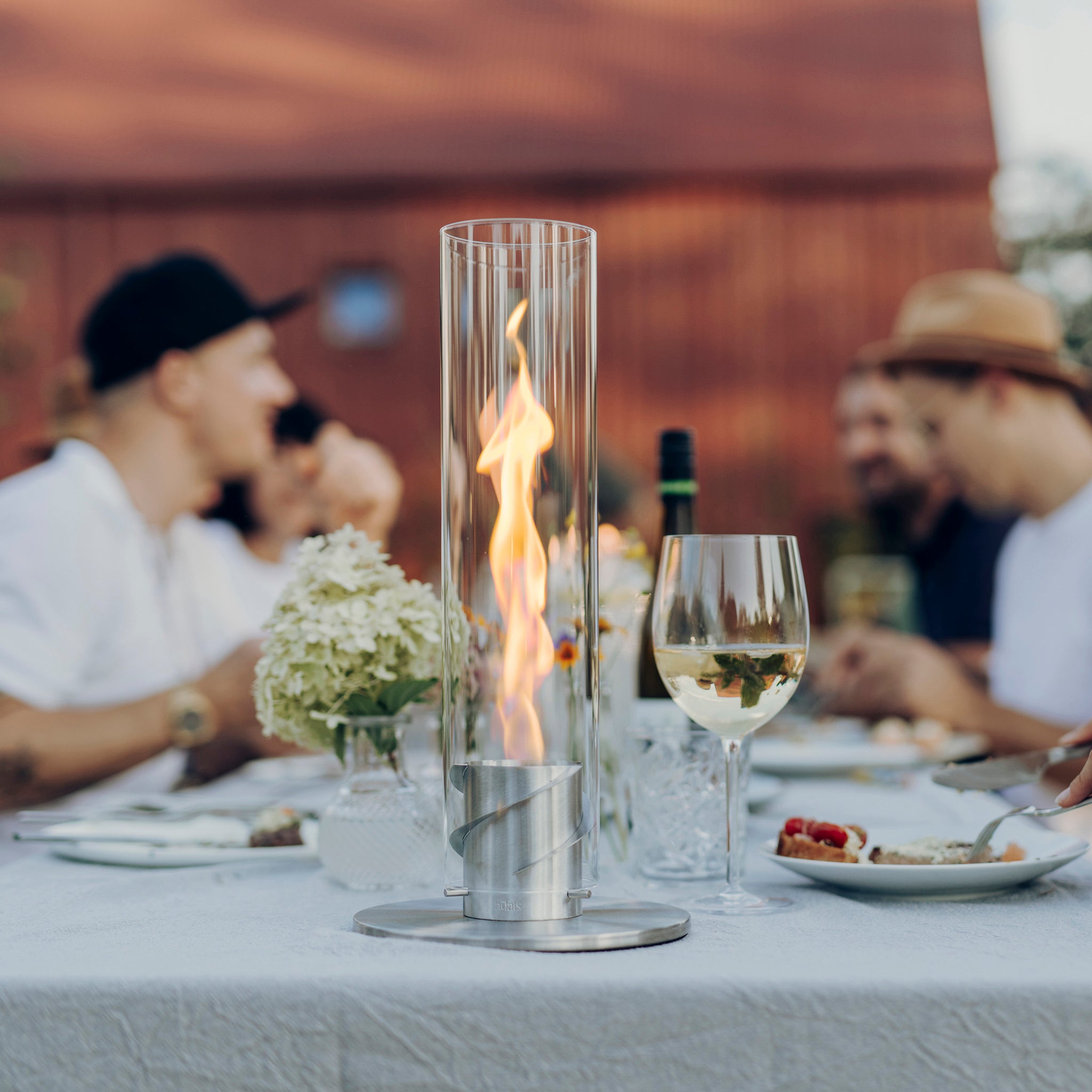 A group of people enjoying an outdoor meal, with a Höfats SPIN 900 Tabletop Fireplace Silver centerpiece in a decorative glass cylinder and food on a white tablecloth.