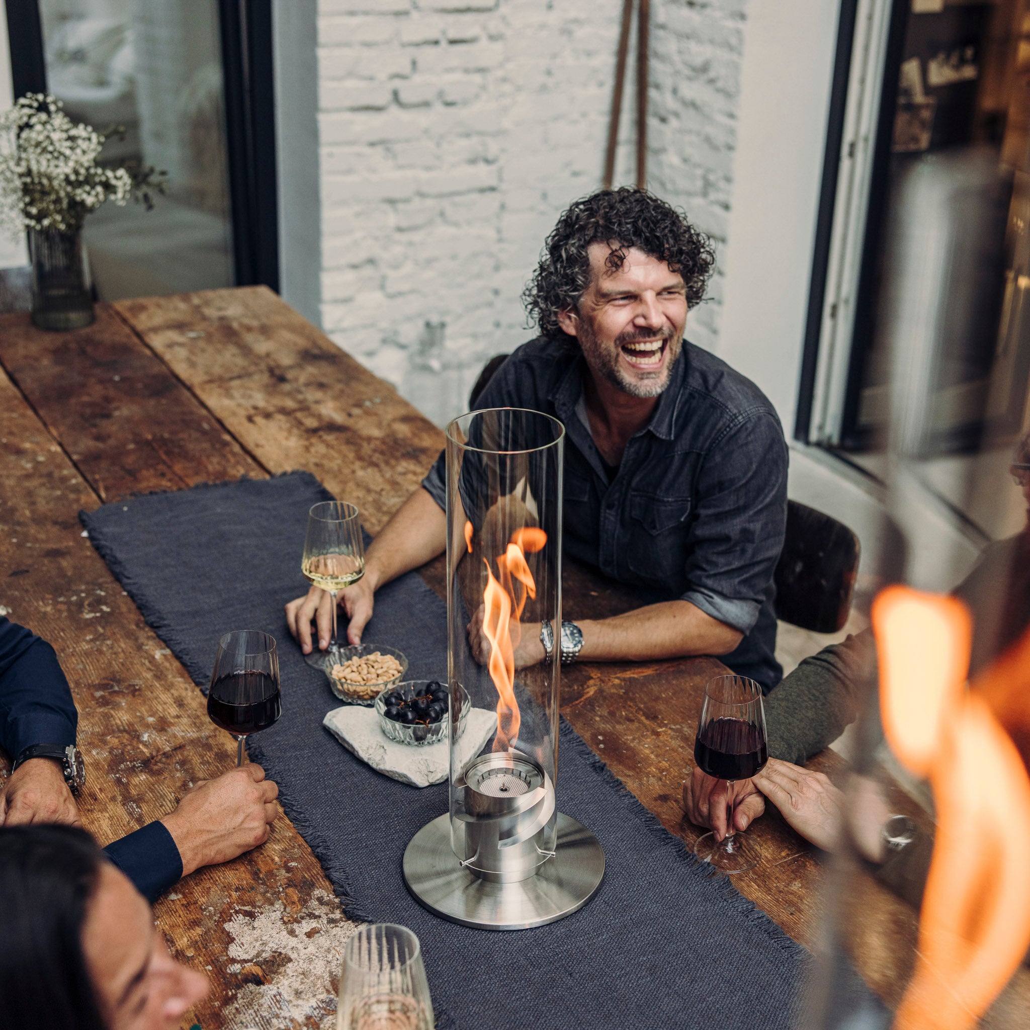 A group of people enjoying drinks around a table with the Höfats SPIN 1200 Tabletop Fireplace Silver casting its warm glow, smiles and laughter all around.