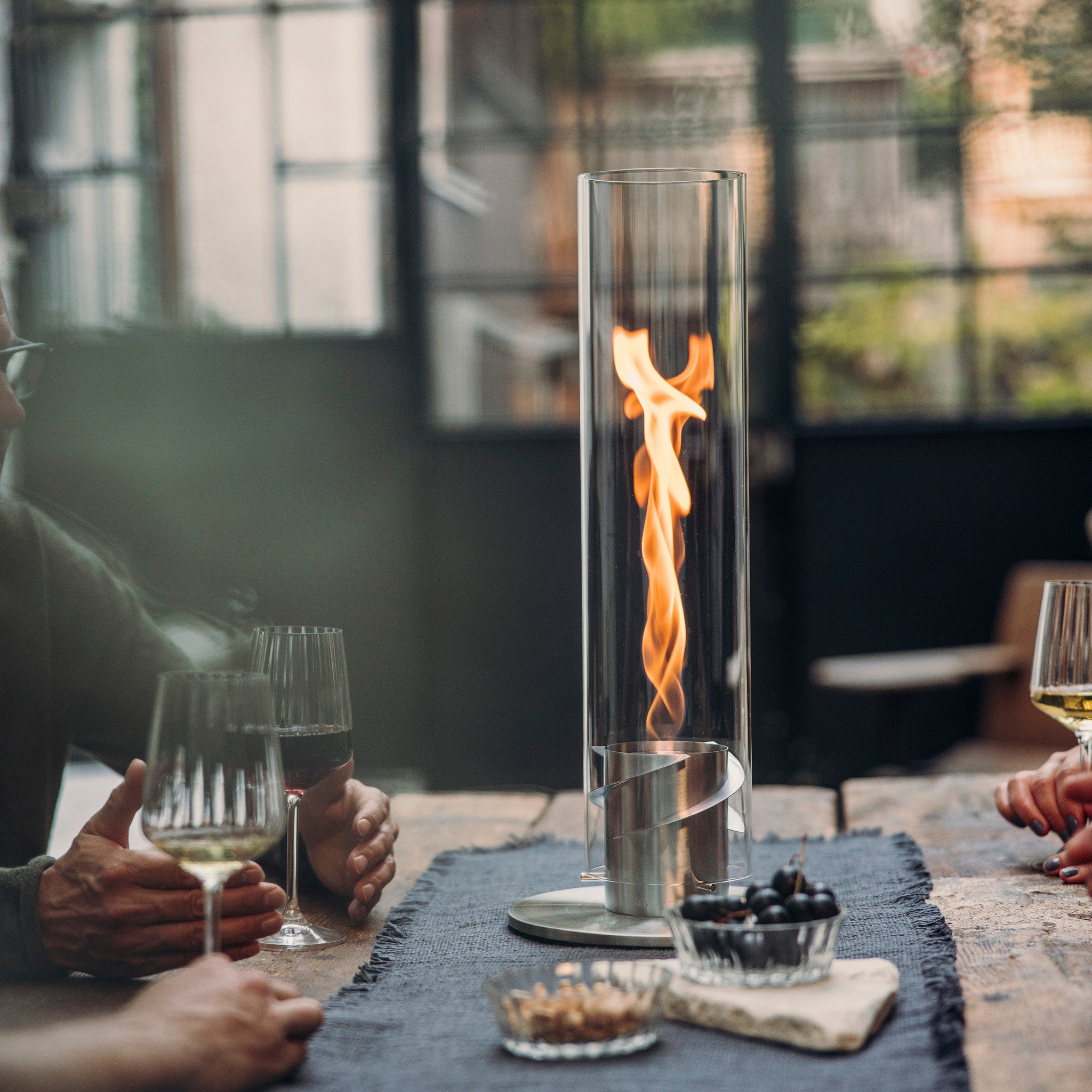 People enjoying drinks around a table with a tall Höfats SPIN 1200 Tabletop Fireplace Silver as a centerpiece, its bioethanol flame dancing elegantly.