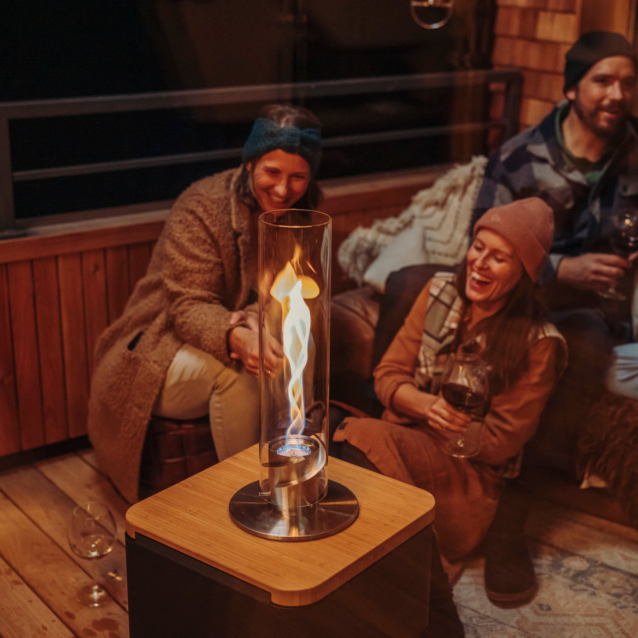 Three people sit around a cozy outdoor fireplace with a Höfats SPIN 1200 Tabletop Fireplace in Silver, sipping wine and enjoying a relaxed evening. The decorative glass cylinder of the bioethanol flame adds a warm and inviting ambiance.