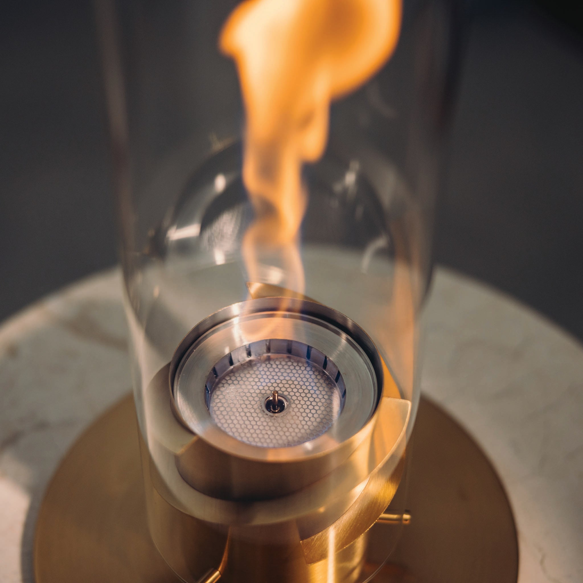 Close-up of the Höfats SPIN 1200 Tabletop Fireplace Silver, featuring a bioethanol flame inside a decorative glass cylinder, set on a brass base.