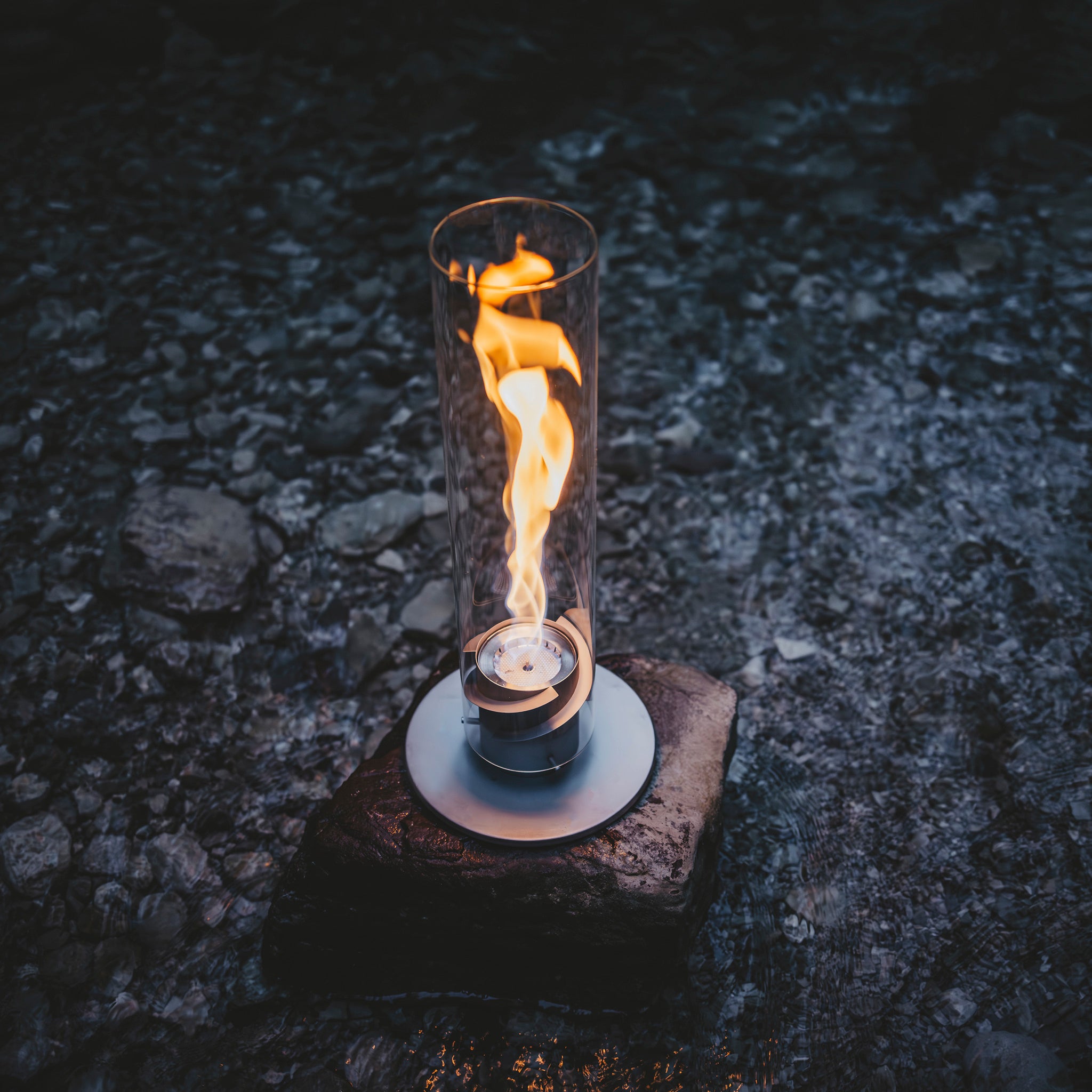 A decorative glass cylinder lantern with a bioethanol flame inside, the Höfats SPIN 1200 Tabletop Fireplace Grey is elegantly placed on a rock surrounded by water.