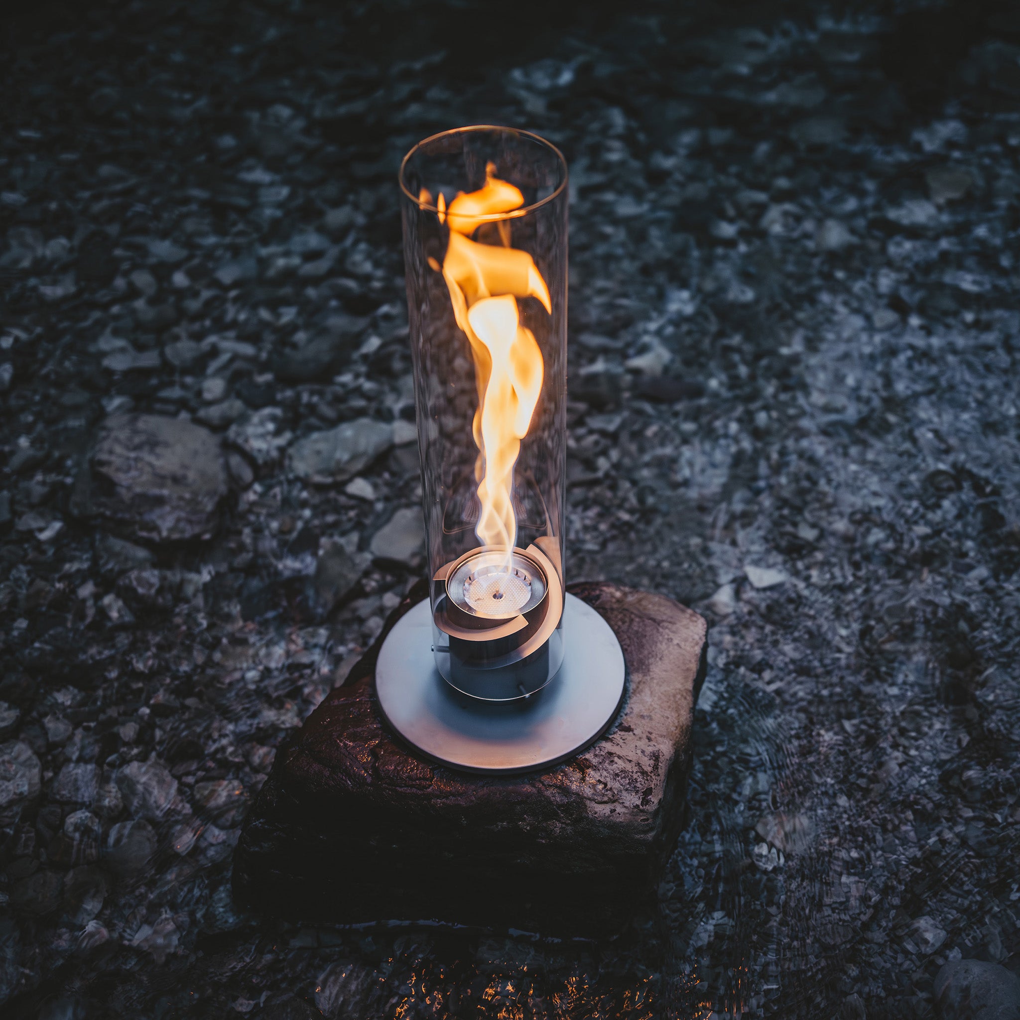 A bioethanol flame inside a decorative glass cylinder beautifully illuminates a stone in the dark, rocky setting, reminiscent of the elegant design of the Höfats SPIN 900 Tabletop Fireplace Grey.