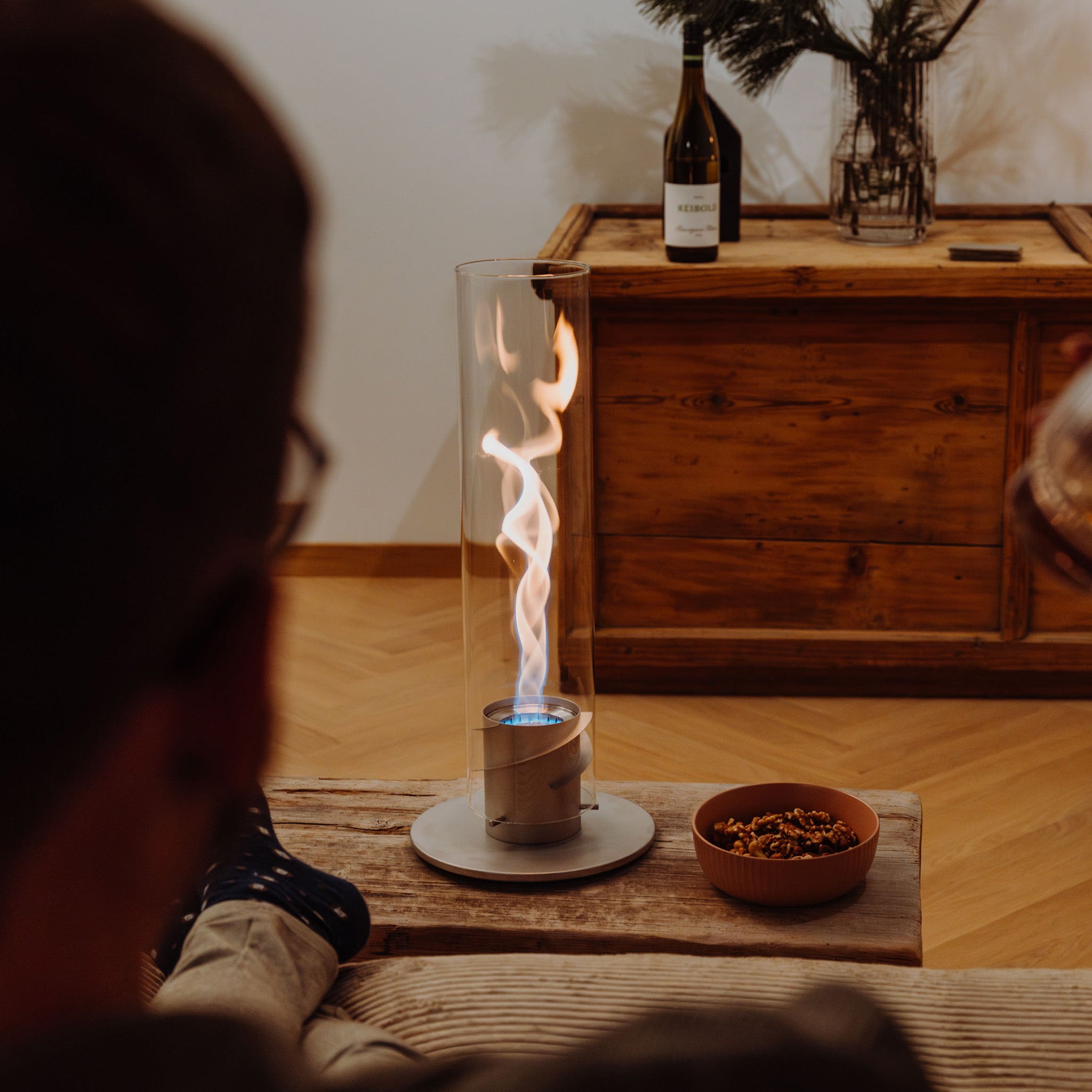 Unwinding with wine and nuts becomes even more enjoyable in a cozy living room setting when you're relaxing by the Höfats SPIN 900 Tabletop Fireplace Grey, encased in its decorative glass cylinder, creating a perfect warm campfire atmosphere.