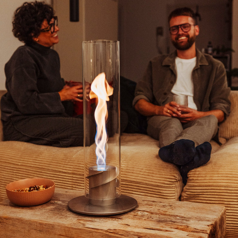 Two people relaxing on a couch with drinks, a Höfats SPIN 1200 Tabletop Fireplace in grey featuring a decorative glass cylinder producing a mesmerizing bioethanol flame, and a bowl of snacks on the rustic wooden table.