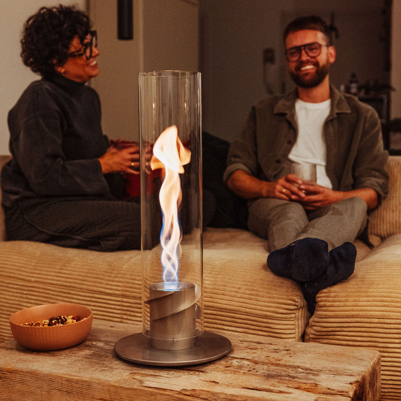 Two people sitting on a couch, smiling, as the Höfats SPIN 900 Tabletop Fireplace in Grey, with its decorative glass cylinder, creates a cozy campfire atmosphere. A bowl of snacks rests on the wooden table, adding to the warmth and charm of this bioethanol-fueled scene.