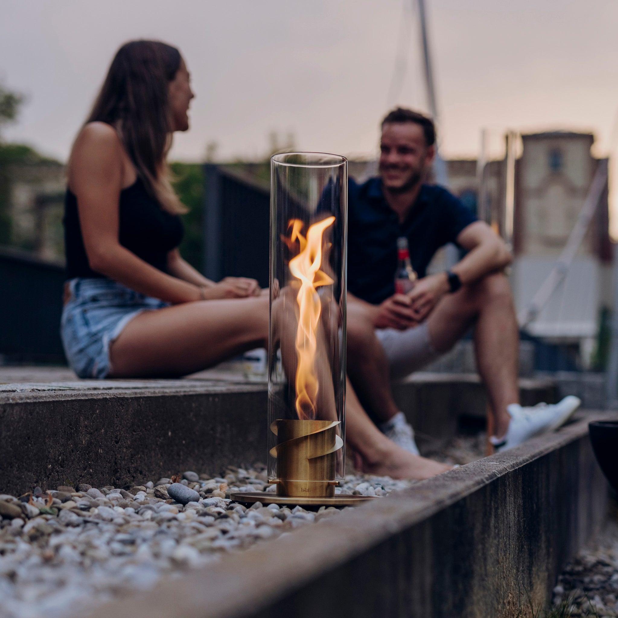 Two people sit on concrete steps with a modern, glass-encased Höfats SPIN 90 Table Top Fire in Gold in the foreground.