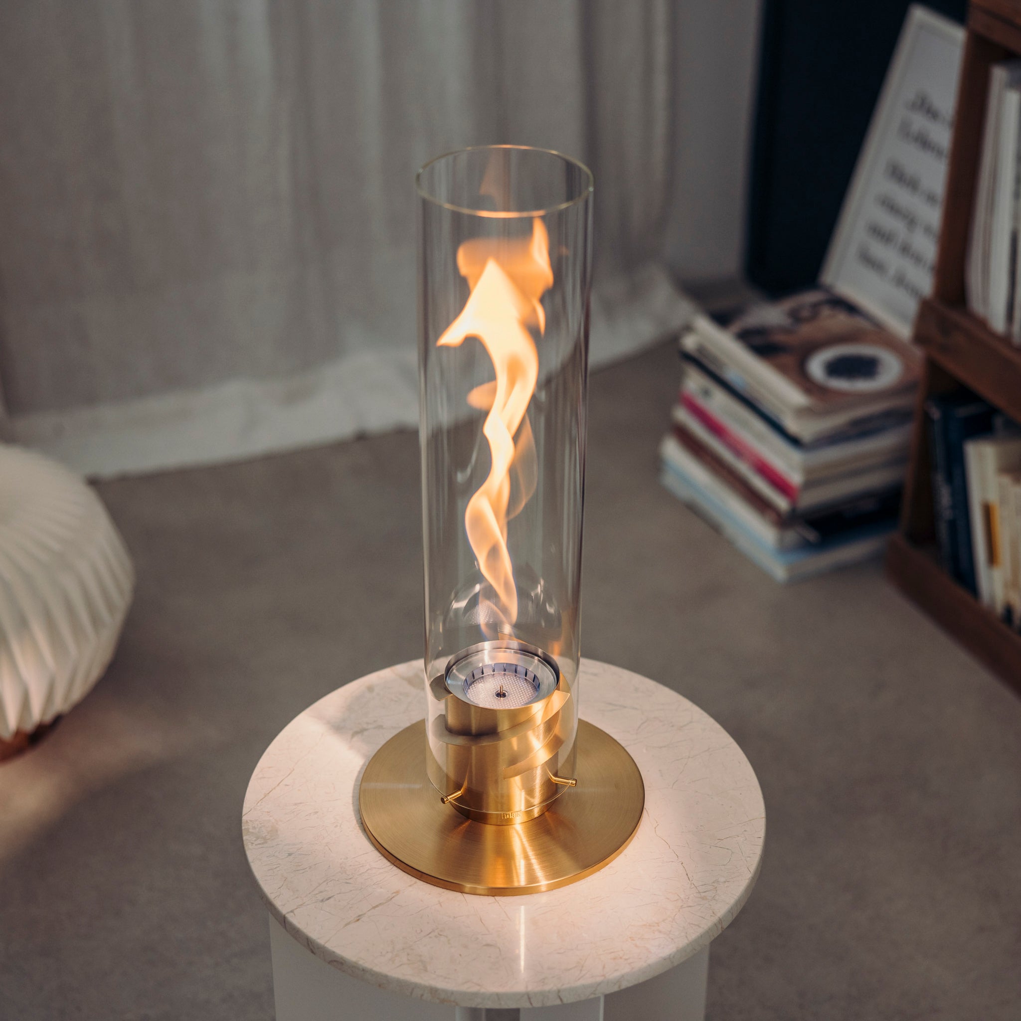 A small tabletop fireplace featuring a decorative glass cylinder with visible bioethanol flames sits on a white surface, exemplifying the elegant design of the Höfats SPIN 1200 Tabletop Fireplace Gold.