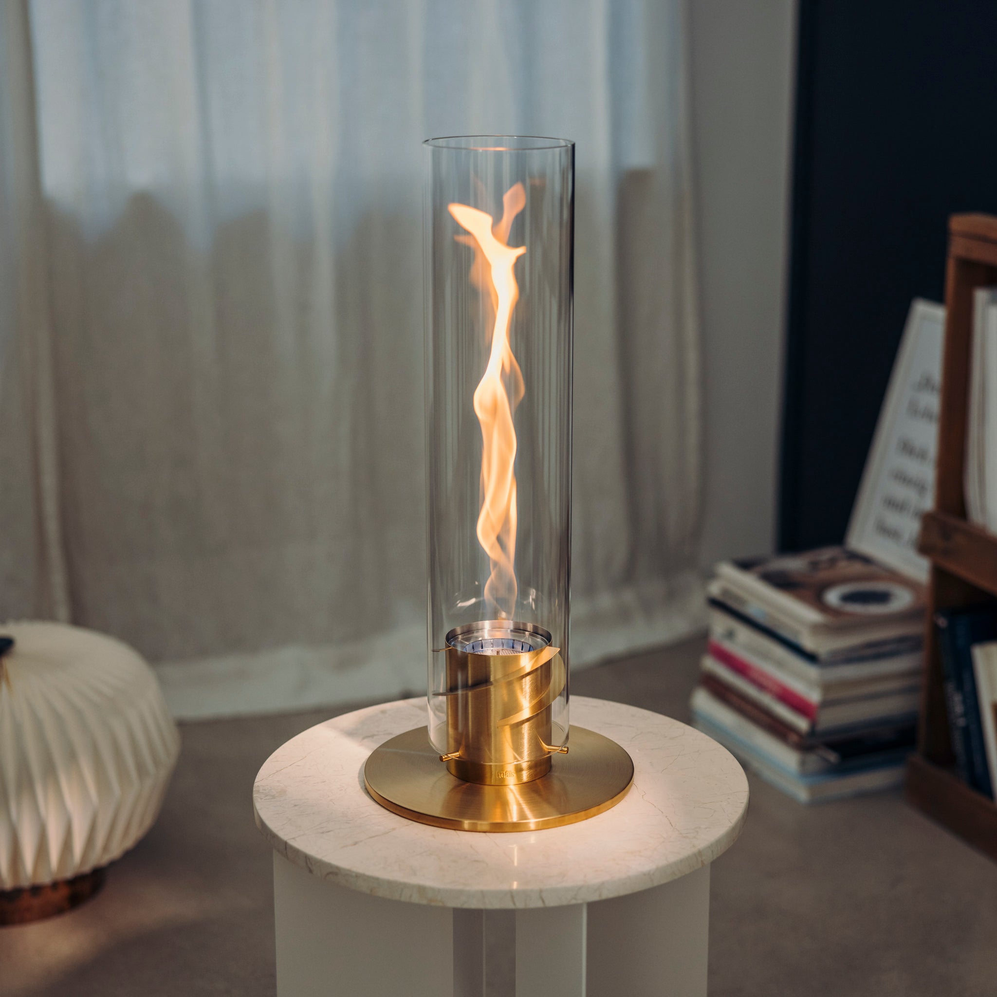 A small, elegant tabletop fire feature with a gold base and a decorative glass cylinder, sitting on a round marble table indoors. The Höfats SPIN 1200 Tabletop Fireplace Gold creates a mesmerizing ambiance perfect for any home decor.