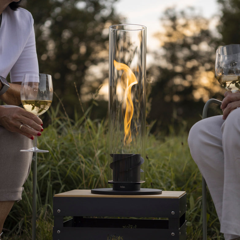 Two people sit outdoors, holding wine glasses, with a Höfats SPIN 90 Table Top Fire in Black containing a bioethanol flame on the table between them.