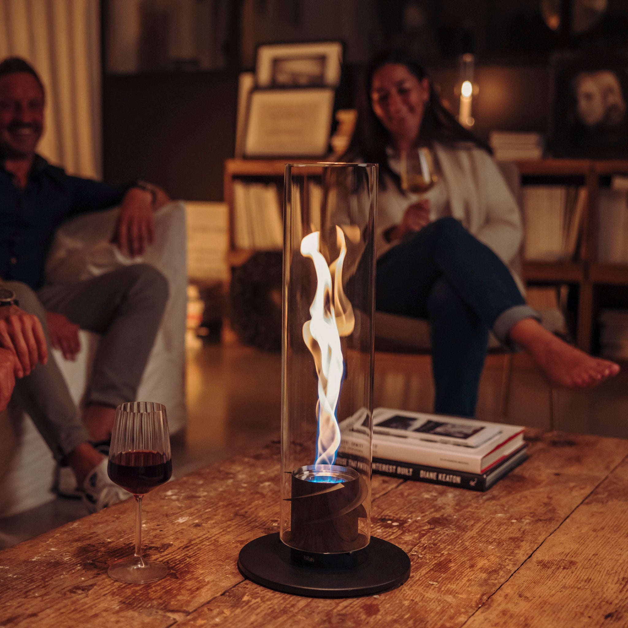 A cozy indoor scene with a Höfats SPIN 1200 Tabletop Fireplace Black on a wooden table, surrounded by relaxed individuals enjoying wine, as the decorative fire adds warmth and charm.