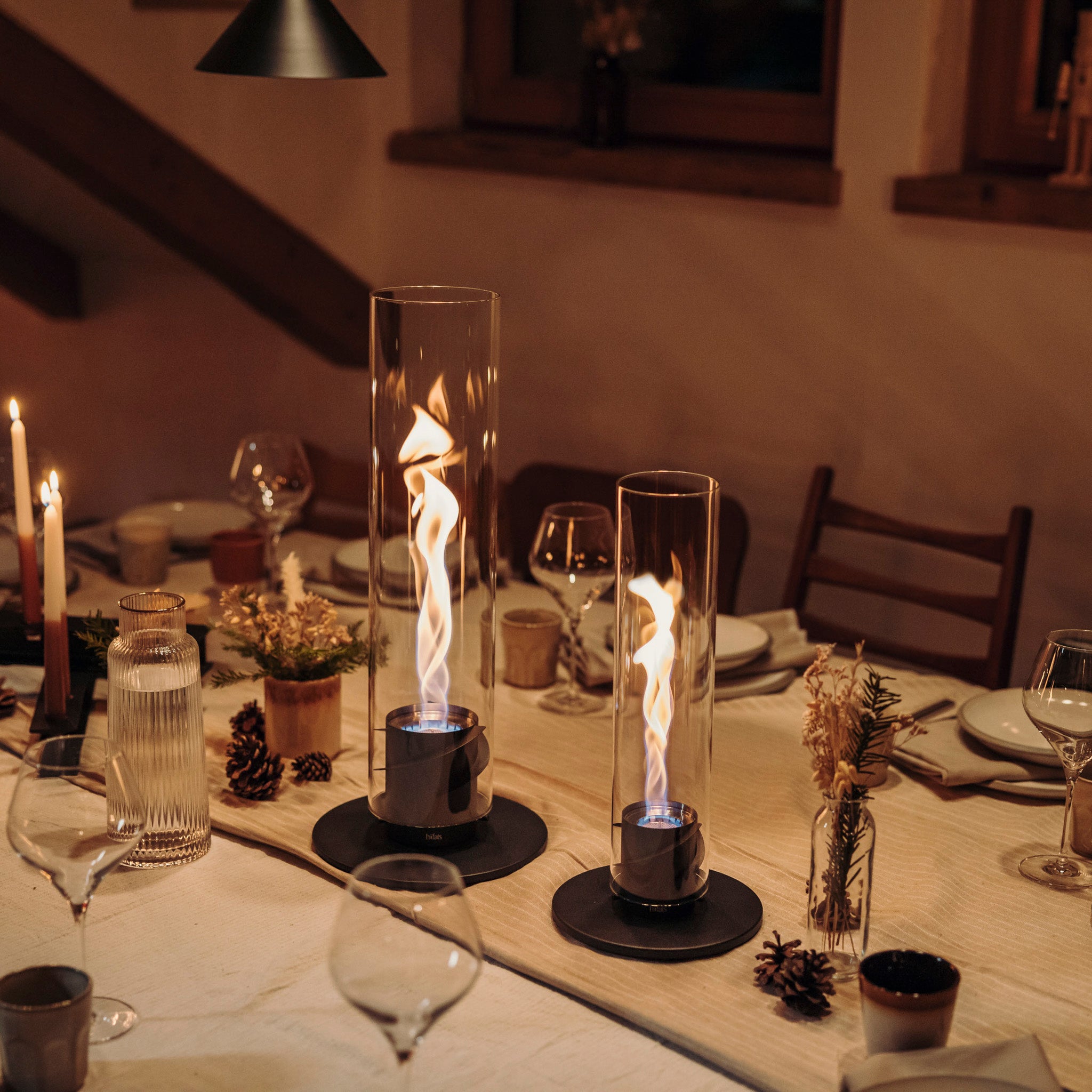 A cozy dining table with candles, wine glasses, and two Höfats SPIN 1200 Tabletop Fireplaces in Black featuring bioethanol flames, set for an intimate dinner.