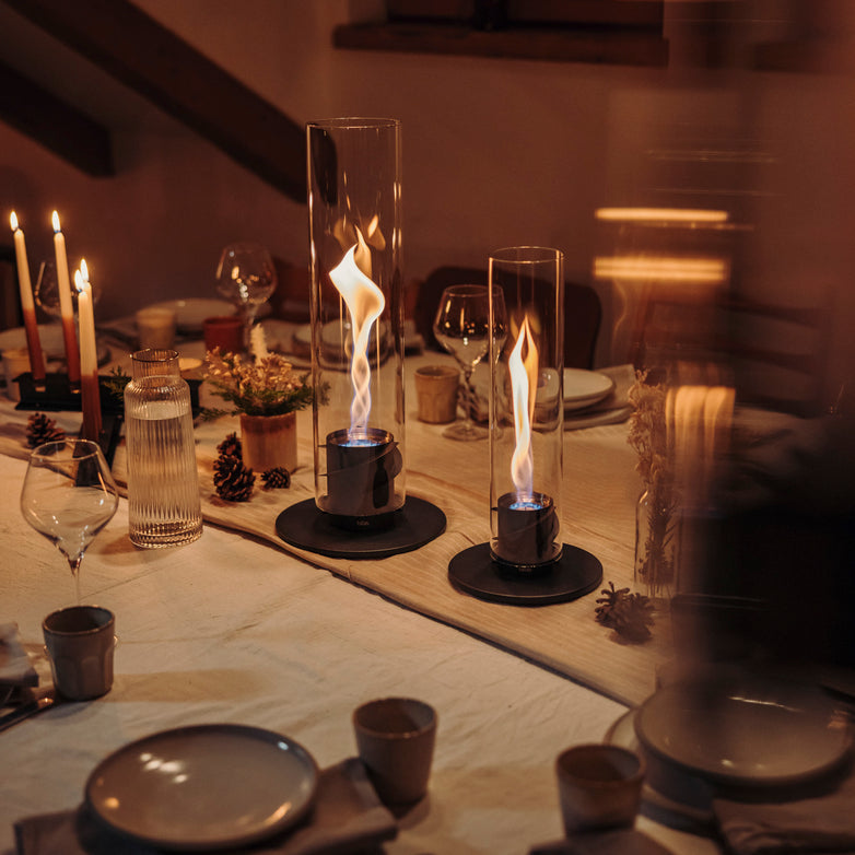 Elegant dinner setting with lit candles, glassware, and two Höfats SPIN 900 Tabletop Fireplaces in black on a white tablecloth. Warm ambiance.