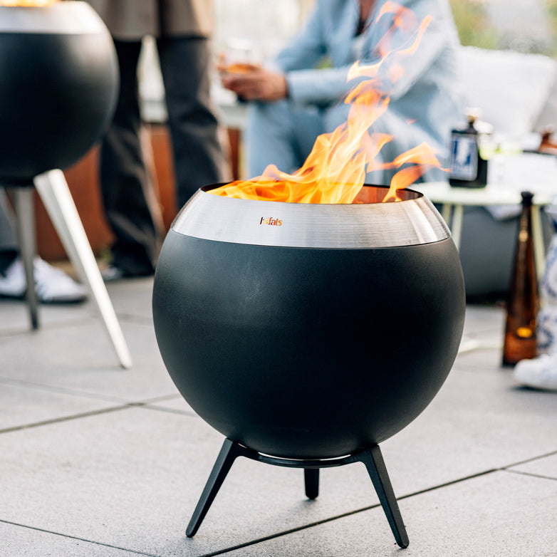 A spherical, black Höfats MOON 45 Fire Bowl with flames rising high on an outdoor patio, people in the background. This environmentally friendly fire bowl utilizes wood gasification for efficient and eco-conscious burning.