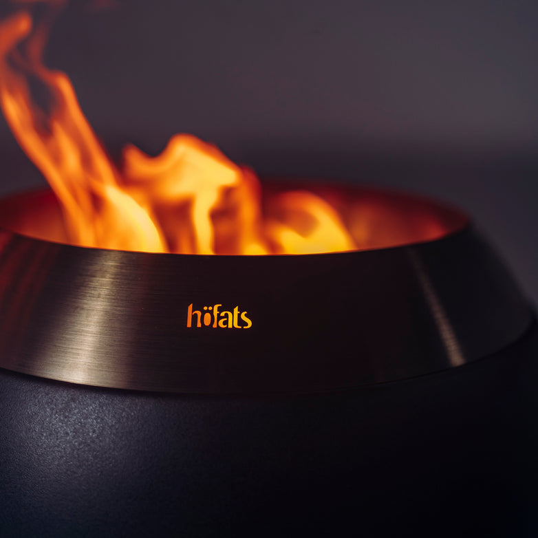 Close-up of a Höfats MOON 45 Fire Bowl with Low Stand, showcasing its blazing flames and the word "höfats" elegantly illuminated on the side. This environmentally friendly fire bowl features sophisticated black MOON fire art, creating an enchanting and sustainable focal point for any outdoor gathering.