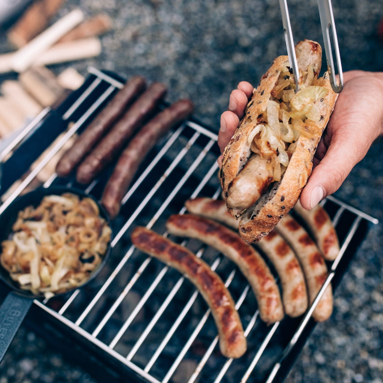 Person holding a grilled sausage in a bun with onions over the Höfats BEER BOX Cooking Grid full of sausages and a pan of grilled onions.