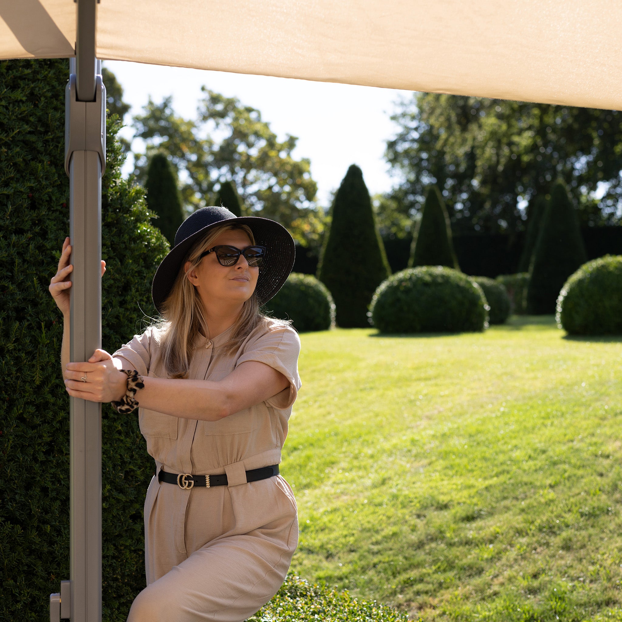 A woman in a beige jumpsuit and black hat stands under the Pallas 4m x 3m Rectangular Cantilever Parasol with LED Lighting in Beige in a manicured garden on a sunny day.