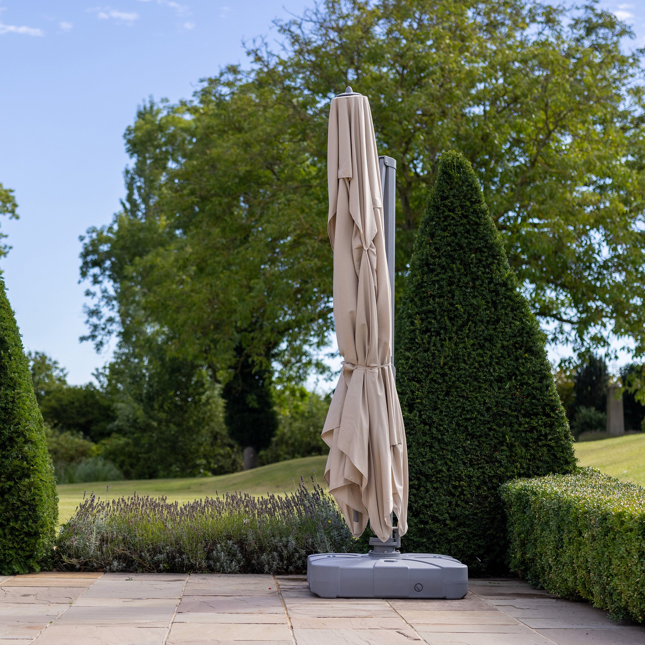 A closed Pallas 4m x 3m Rectangular Cantilever Parasol with LED Lighting in Beige, on a tiled outdoor area surrounded by trimmed hedges and lush green trees, provides garden shade for relaxing afternoons.