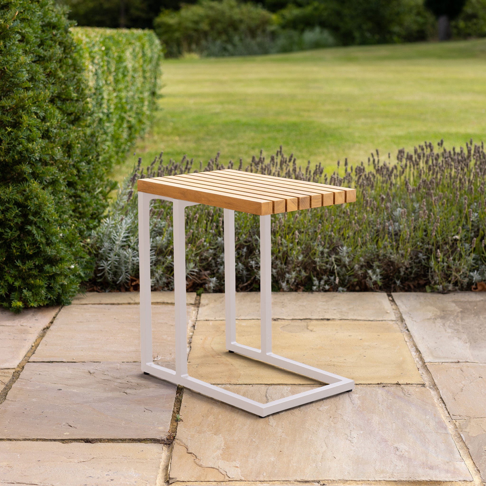 A wooden and metal stool sits elegantly on a stone patio beside a Cube Side Table with a Teak Top in Latte. In the background, a green hedge and lush lawn enhance the serene atmosphere.