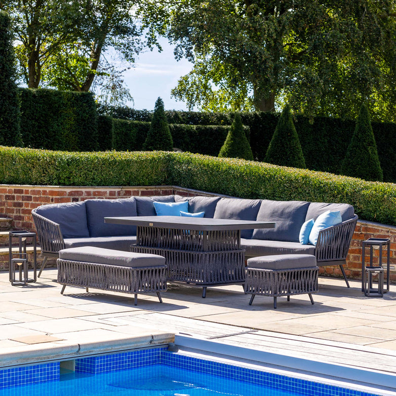 Outdoor patio set featuring the Monterrey Rope Rectangular Corner Dining Set with Rising Table in Grey (Left Hand) by a pool, surrounded by hedges and trees. This Harbour Lifestyle collection includes a gray rattan corner sofa, coffee table, and ottomans, offering a versatile rising table perfect for entertaining.