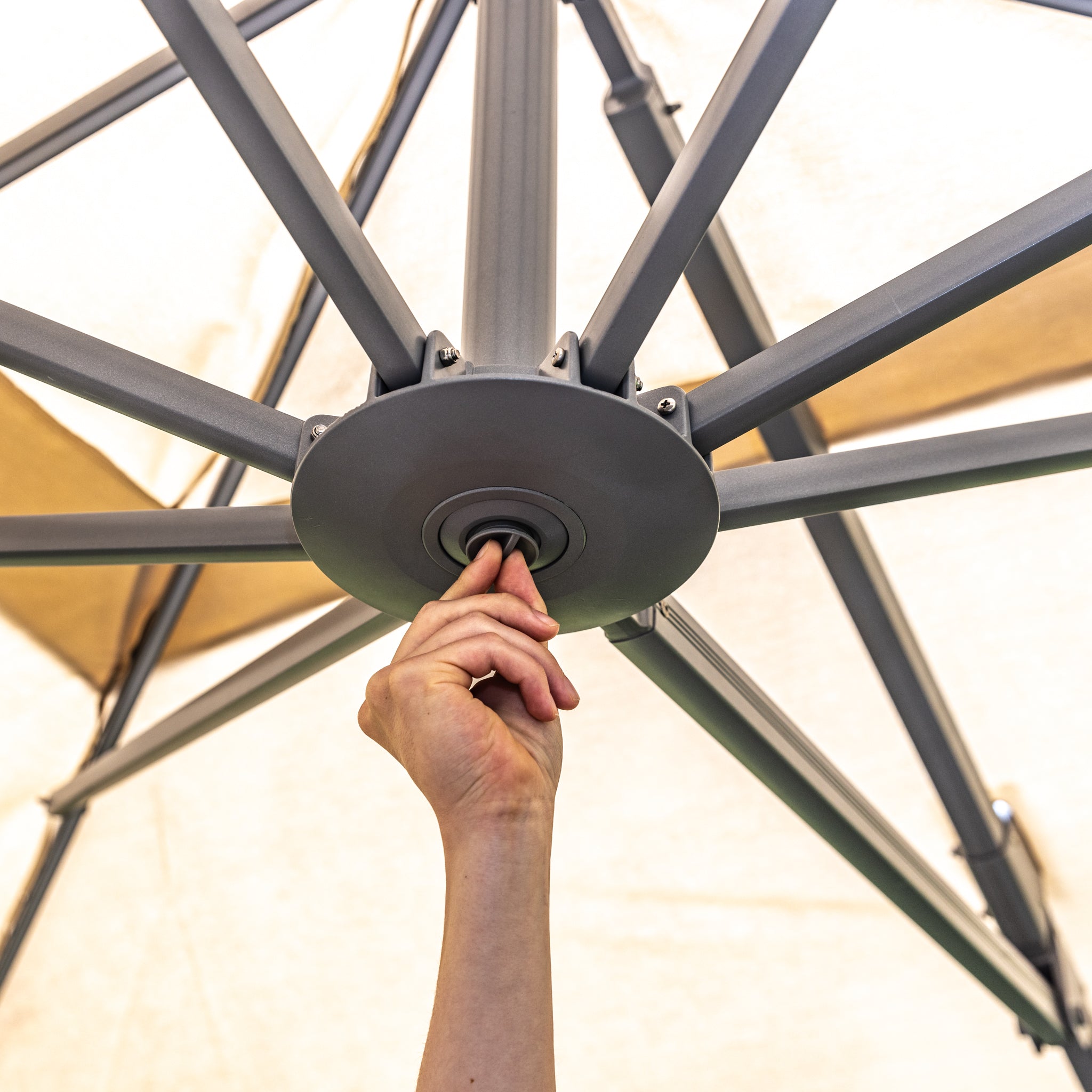 A person adjusting the central mechanism of a Pallas 4m x 3m Rectangular Cantilever Parasol with LED Lighting in Beige from underneath the opened canopy, ensuring perfect garden shade for a sunny afternoon.