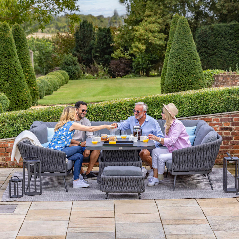 Four people seated around an outdoor table with the Palma U-Shape Rope Corner Dining Set with Rising Table in Grey, enjoying drinks and food in a garden setting with lush greenery.