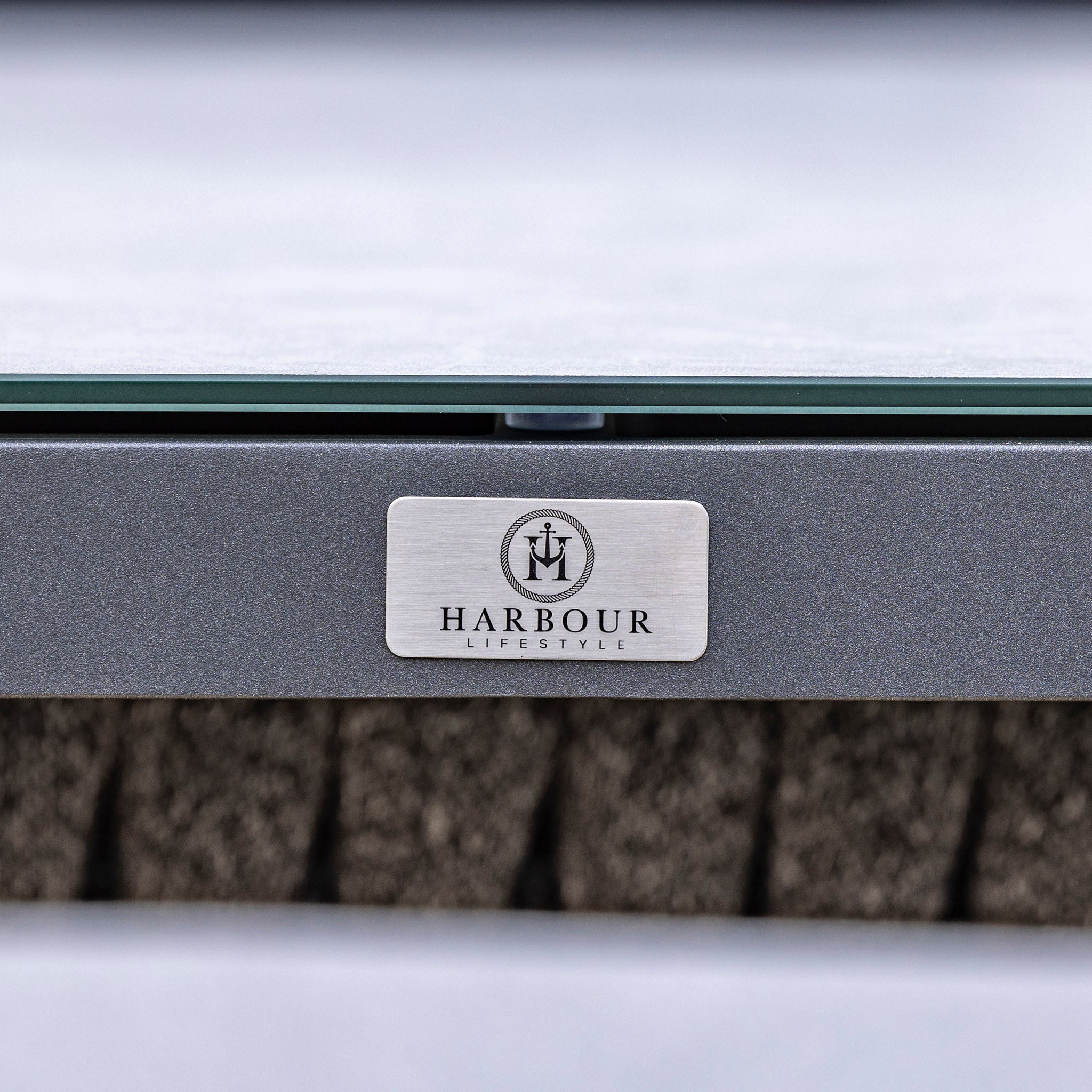 Close-up view of a metallic logo plate with "Harbour Lifestyle" branding on a piece of outdoor furniture, featuring the Palma Deluxe Square Rope Corner Dining Set with Rising Firepit Table in Grey and its hydrophobic rope weave.