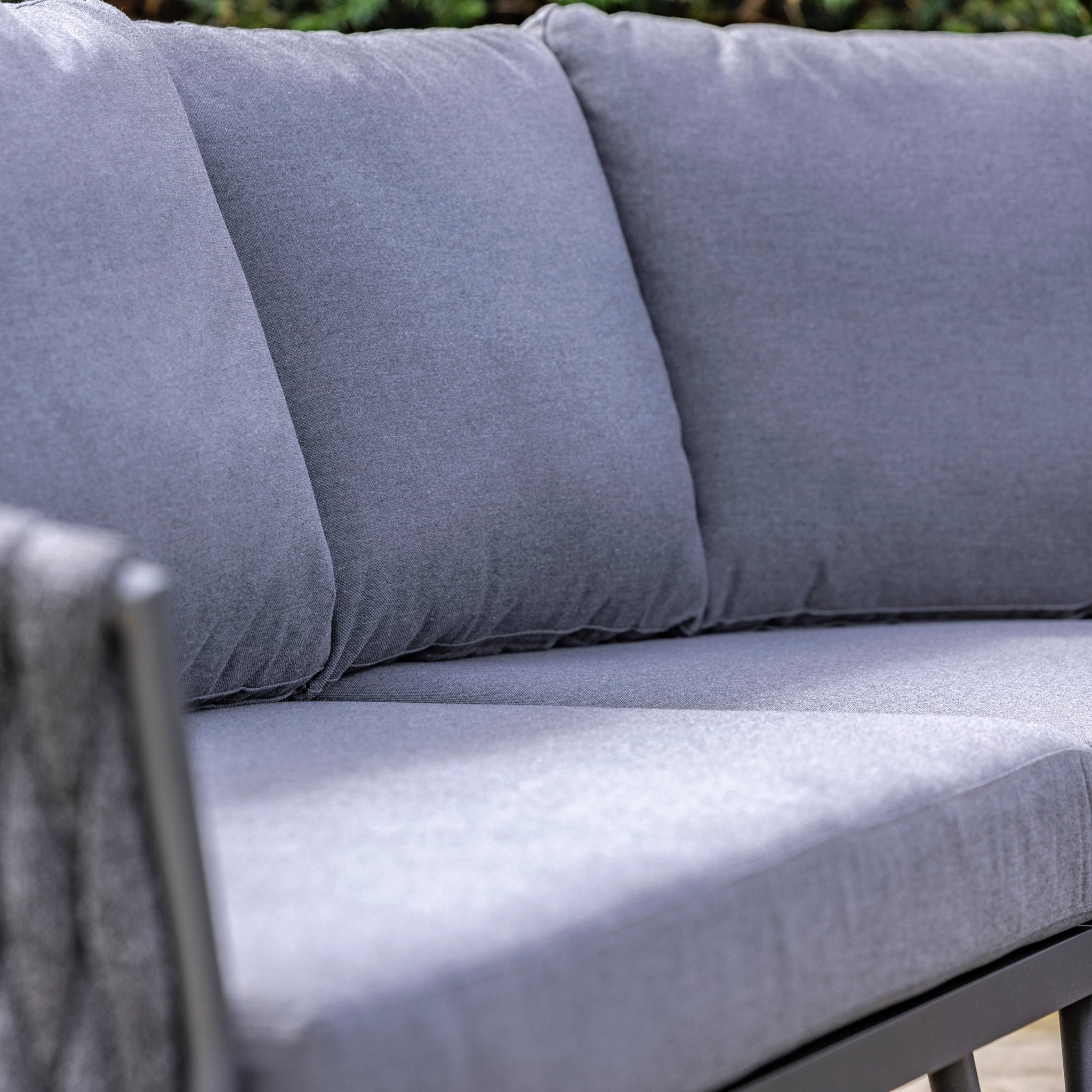 Close-up of a grey outdoor cushioned sofa with an aluminium frame and green foliage in the background, reminiscent of the hydrophobic thick rope weave on the Palma Rope Rectangular Corner Dining Set with Rising Firepit Table in Grey (Left Hand).