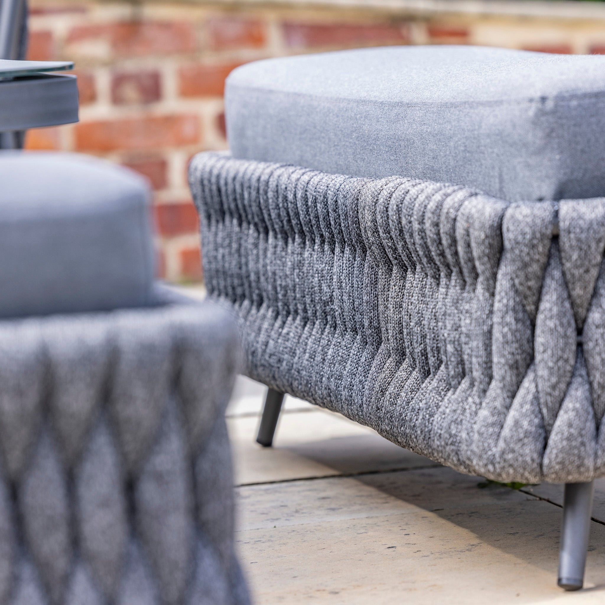 Close-up of a modern, grey outdoor ottoman with a water-repelling thick rope weave base and sleek legs on a patio with a brick background, perfect to pair with the Palma Rope Rectangular Corner Dining Set with Rising Table in Grey (Right Hand).