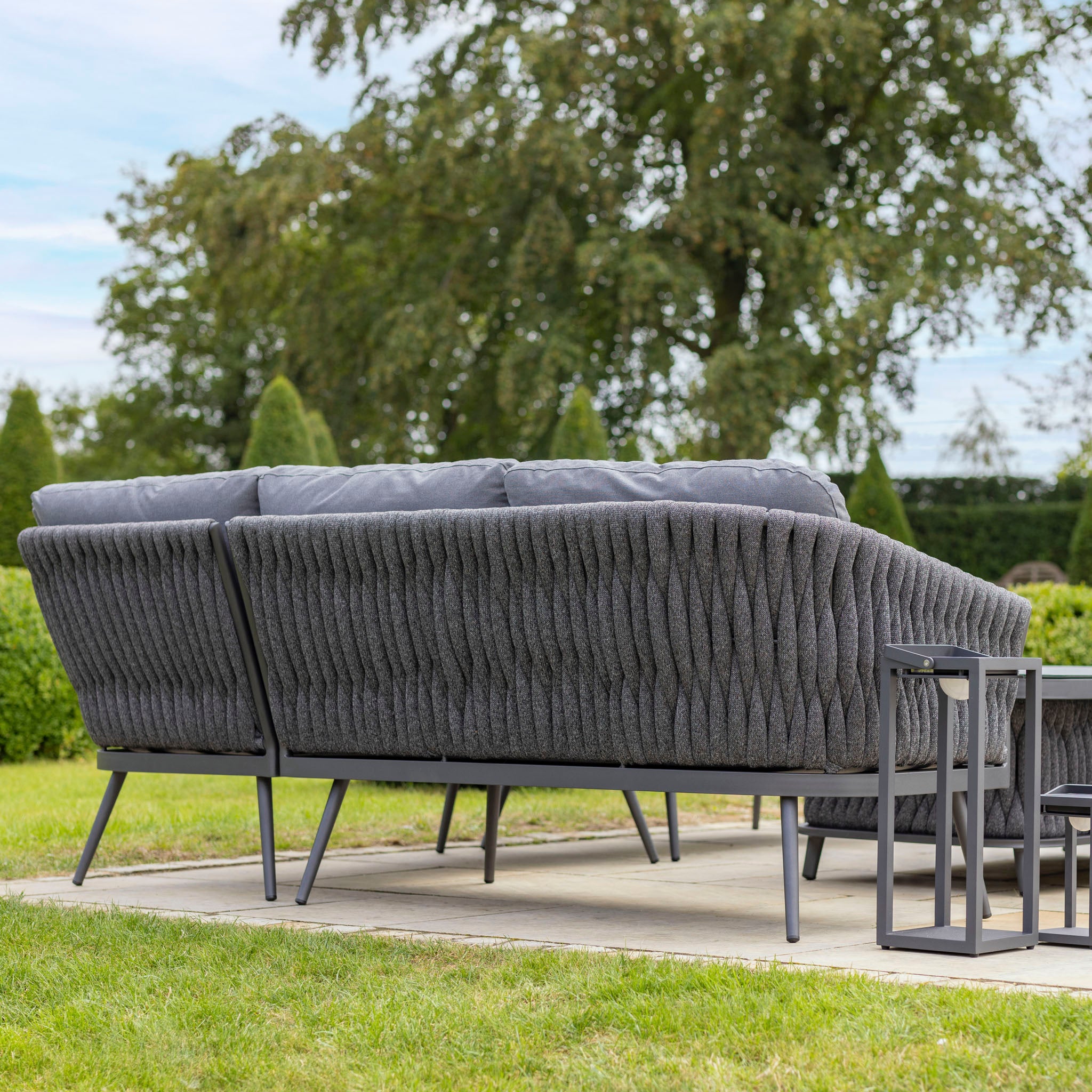 View from the back of the Palma Rope Rectangular Corner Dining Set with Rising Table in Grey (Right Hand), showcasing a modern outdoor patio setup. The dining set includes a dark gray sofa and small tables situated on a concrete slab, all within a garden setting. Its aluminum frames support a water-repelling thick rope weave, offering both durability and style.